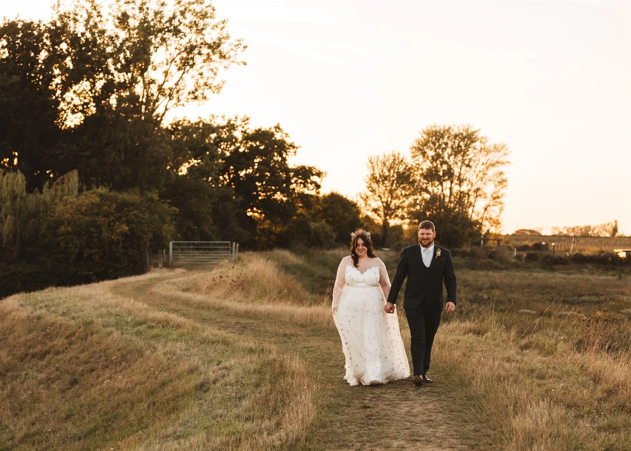 A happy couple walking hand in hand on a grassy path in a rural field during sunset. The woman is wearing a white wedding dress with floral details, and the man is wearing a black suit with a tie.