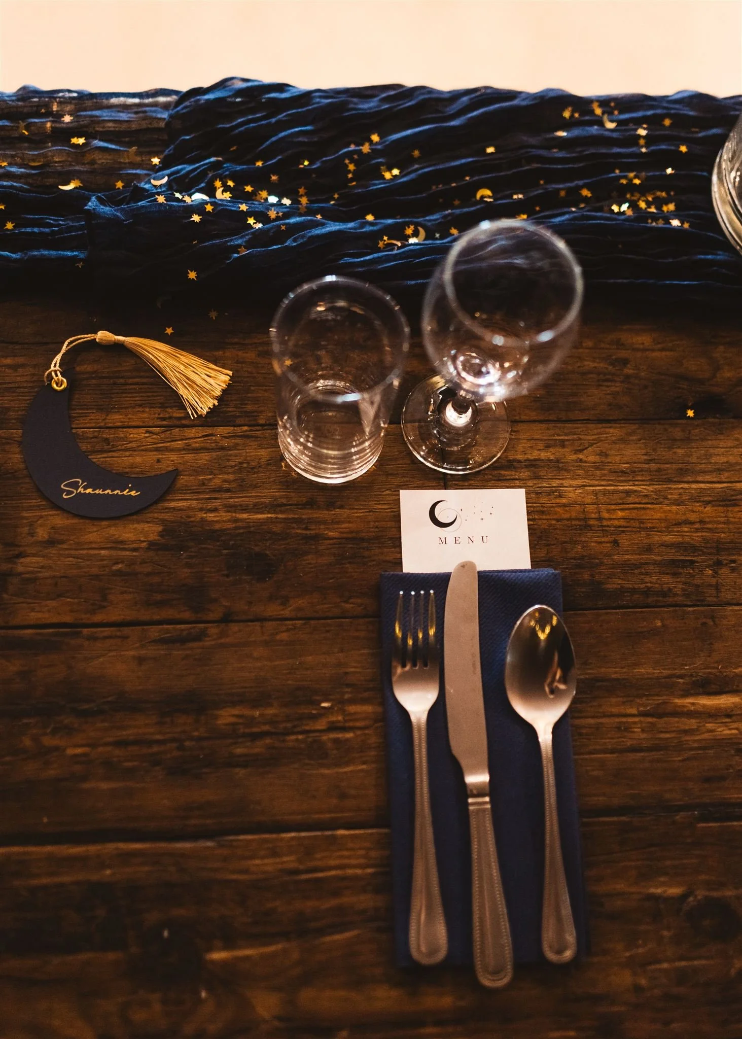 A rustic wooden table set for a dinner with two glasses, a navy blue napkin, and a fork and spoon. There is a small white card with a moon icon and the word 'Menu,' a black name tag with a golden tassel that reads 'Shawnace,' and a navy blue fabric r