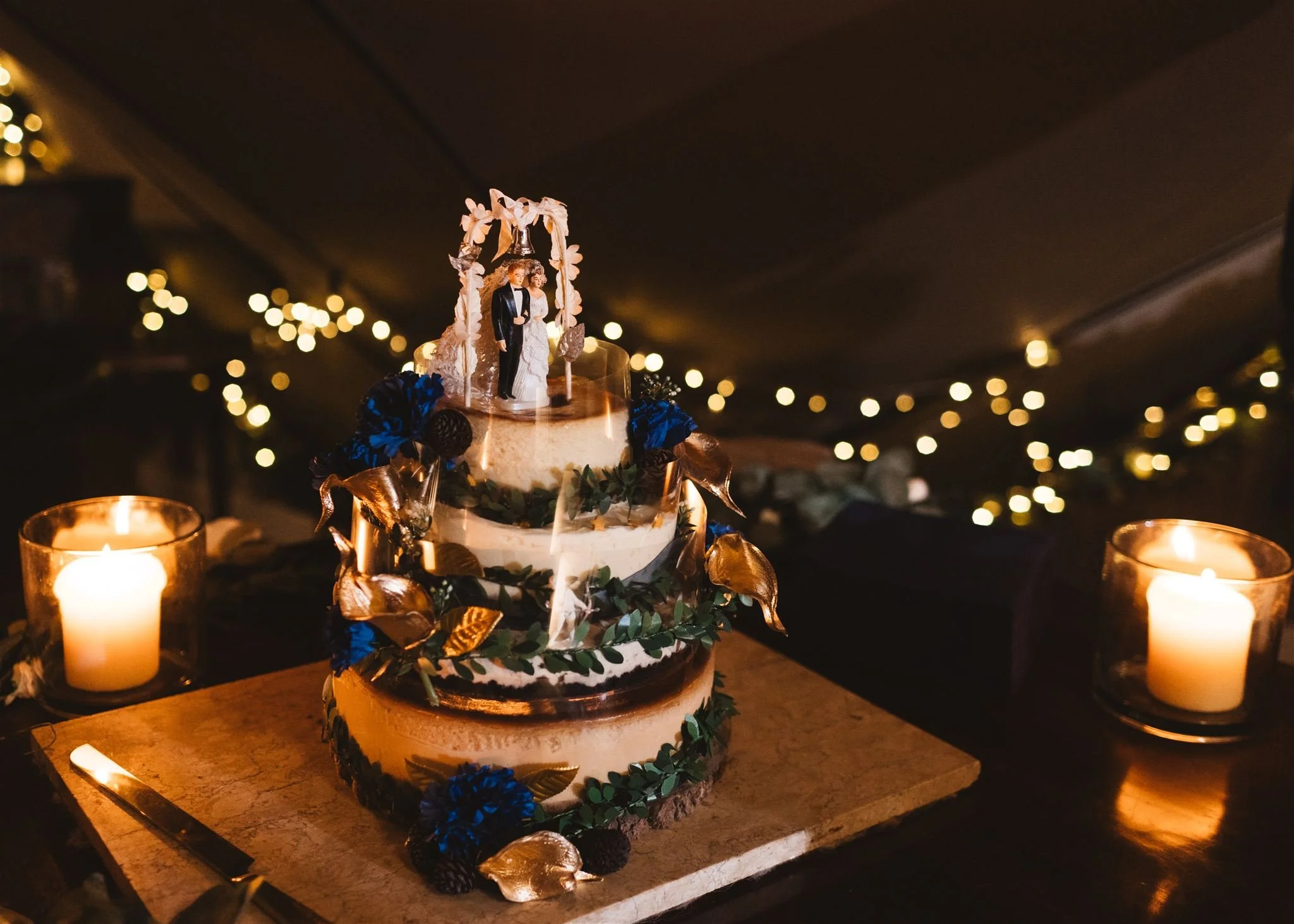 Wedding cake decorated with black, gold, and blue flowers, topped with miniature bride and groom figurines, surrounded by lit candles and string lights in the background.