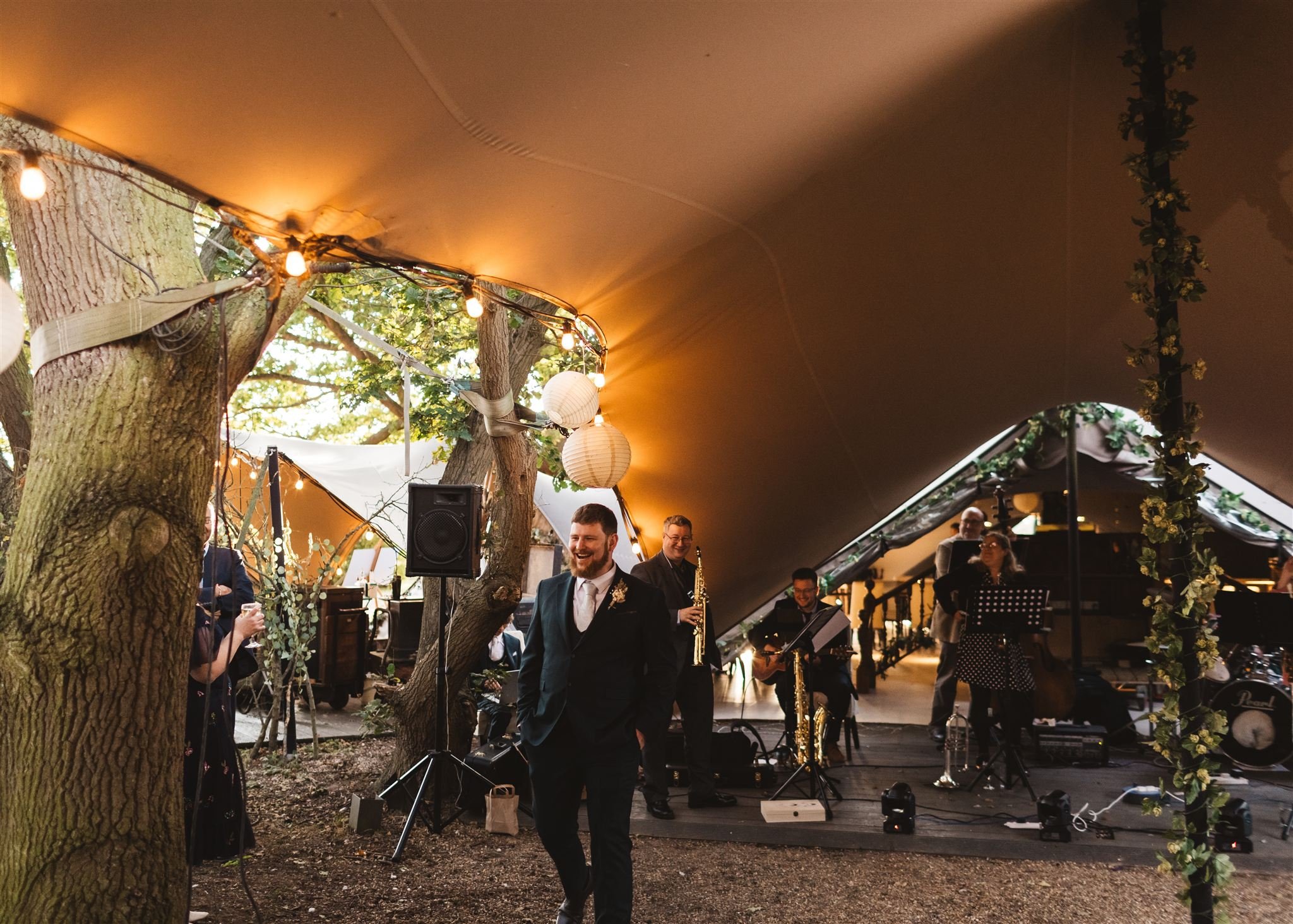 Man in a suit smiling at a wedding reception under a large tent decorated with string lights and paper lanterns, with a live band playing in the background.