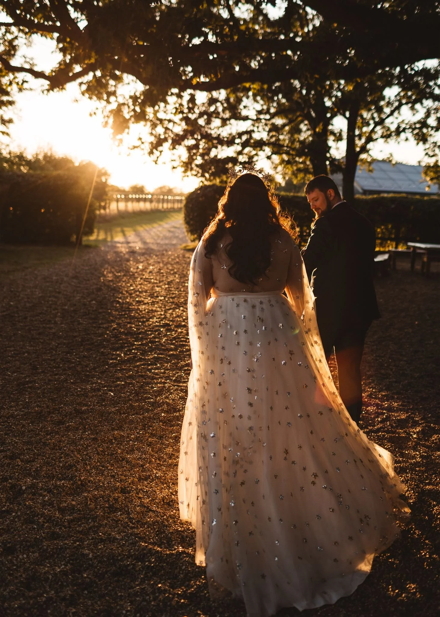 A bride and groom walking outdoors at sunset, with the bride wearing a white gown adorned with sparkling embellishments and the groom in a dark suit, surrounded by trees and a gravel path.