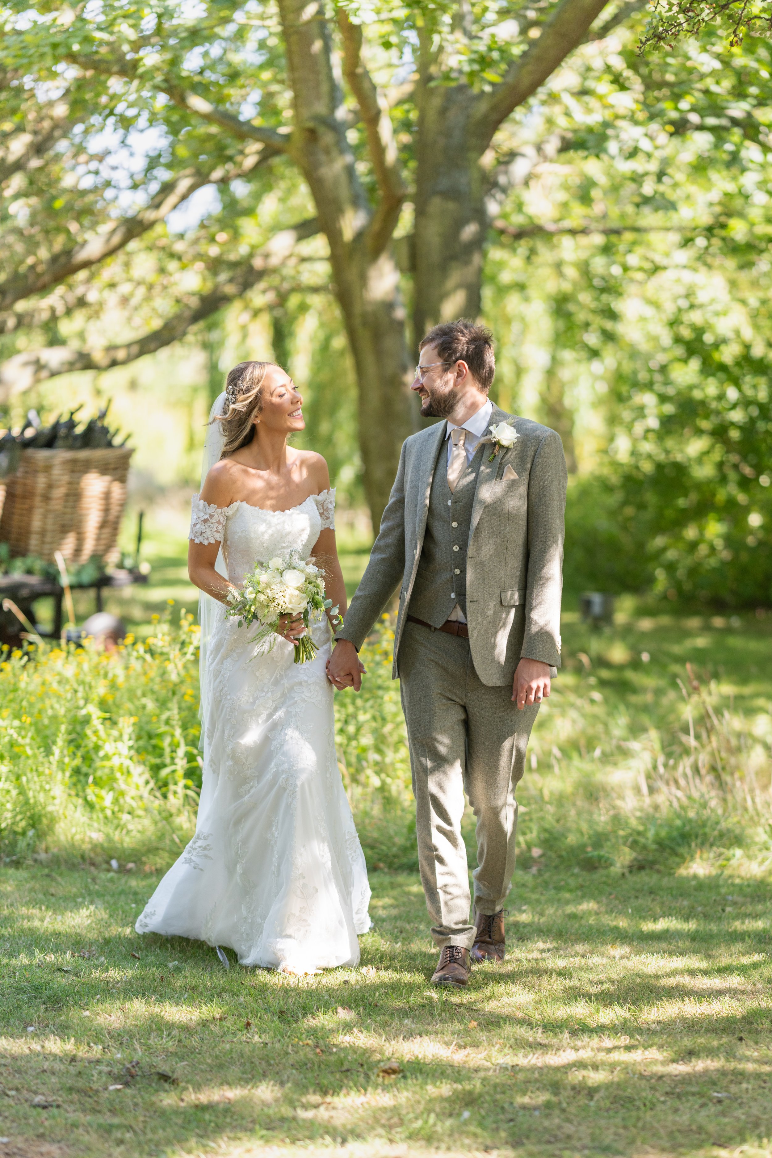 A bride and groom holding hands and walking through a green outdoor setting, smiling at each other on their wedding day.