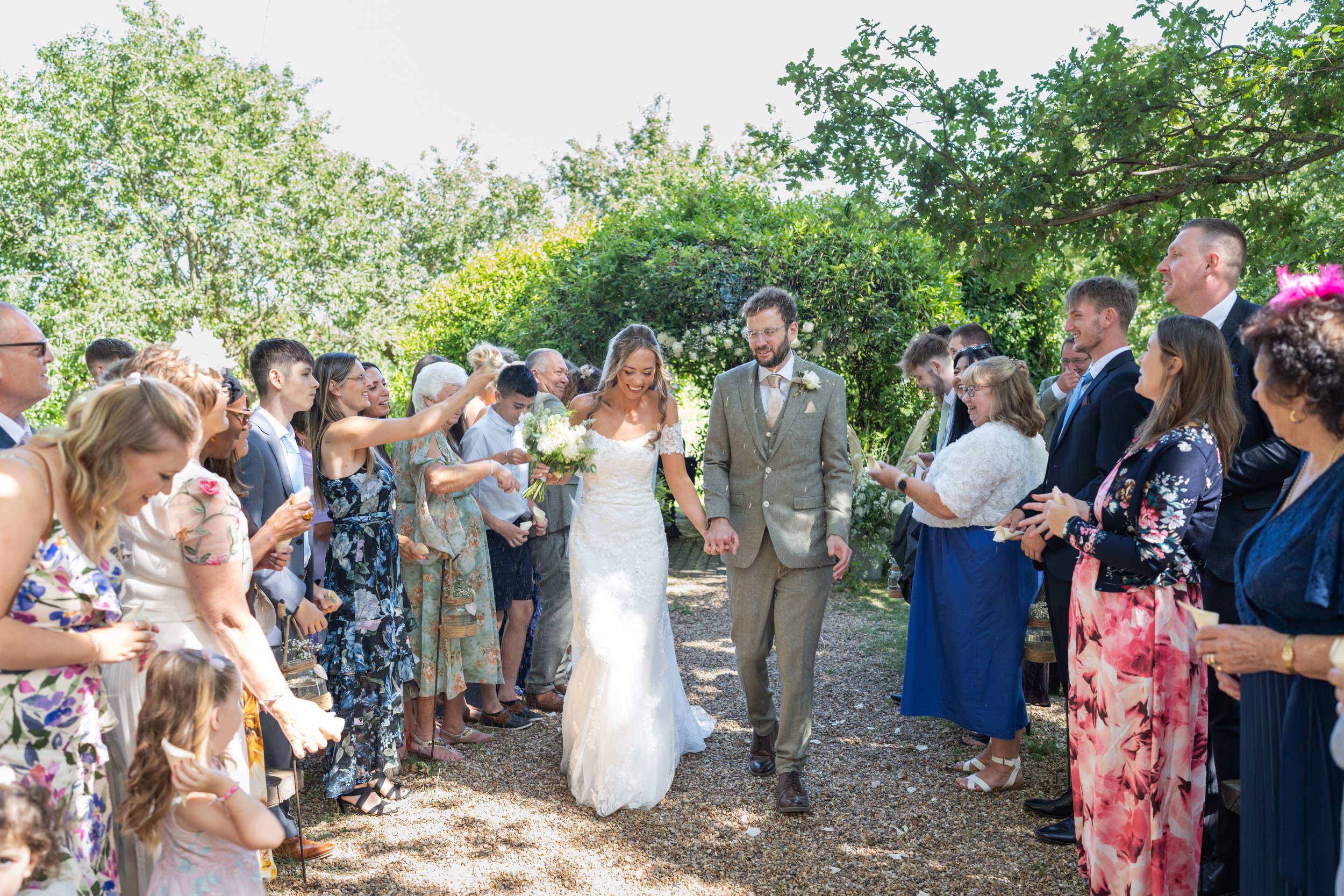 Bride and groom walking hand in hand through a crowd of wedding guests outdoors on a sunny day, with trees and greenery in the background.
