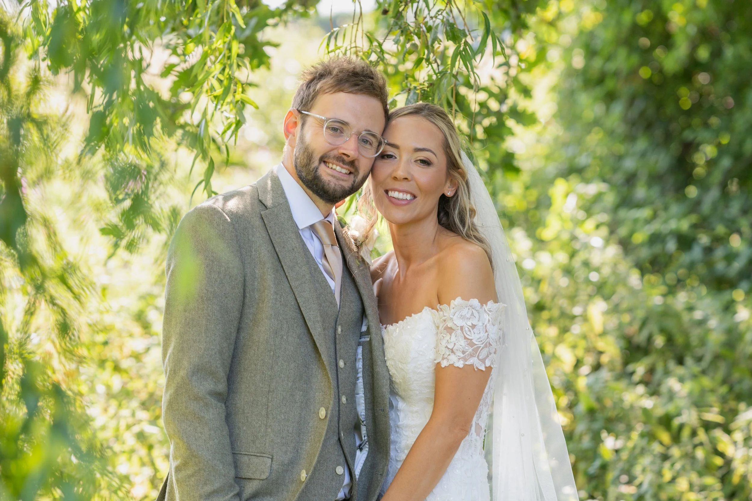 A happy bride and groom in wedding attire posing outdoors among green trees and foliage.