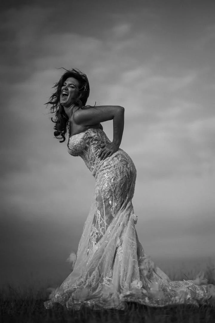 A woman in a lace wedding dress is outdoors, laughing and holding her back, with her hair in motion against a cloudy sky.