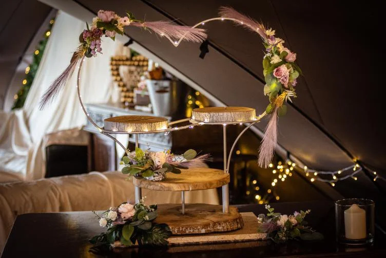 Decorative floral and feather arch with wooden platforms, set on a table with greenery and flowers, in a cozy, festively decorated room with warm lighting.