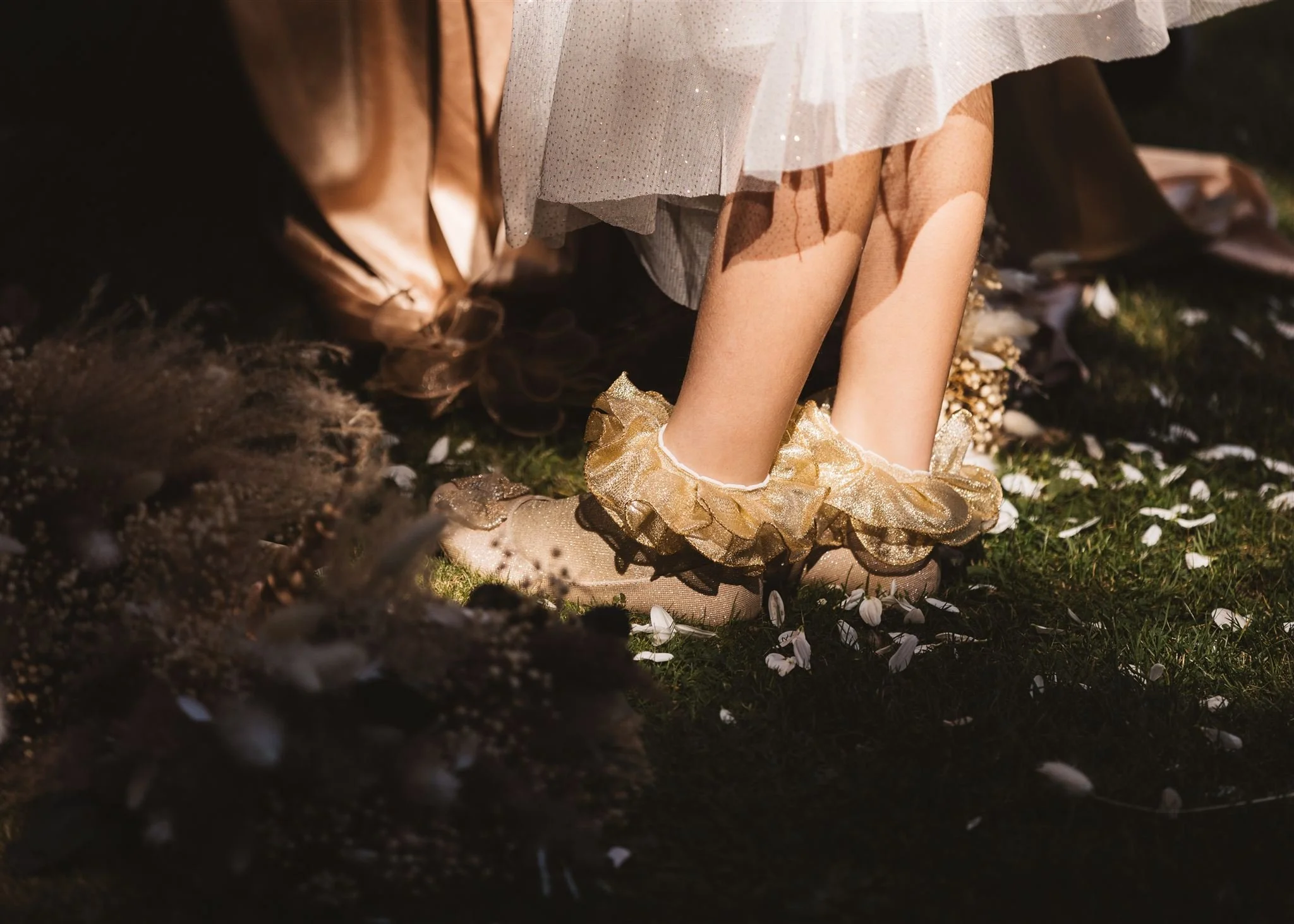 Close-up of a young girl wearing fairy-tale themed shoes with gold and glittery ruffles, standing on grass surrounded by flower petals, part of a costume and dress suggest a fairy-tale or fantasy setting.