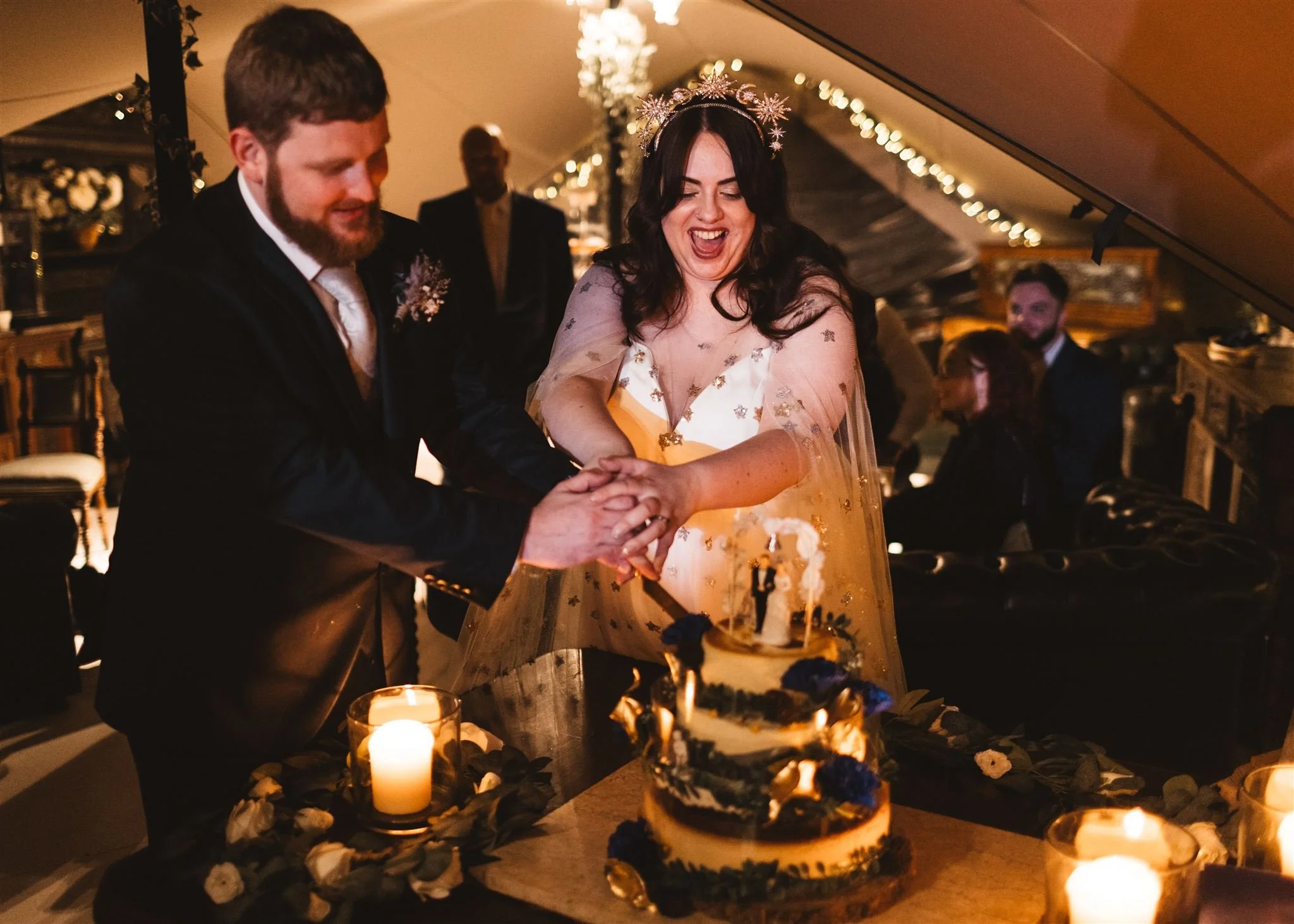 A wedding celebration where a bride and groom are cutting a cake together, with candles and floral decorations on the table in a warmly lit, elegant indoor setting.