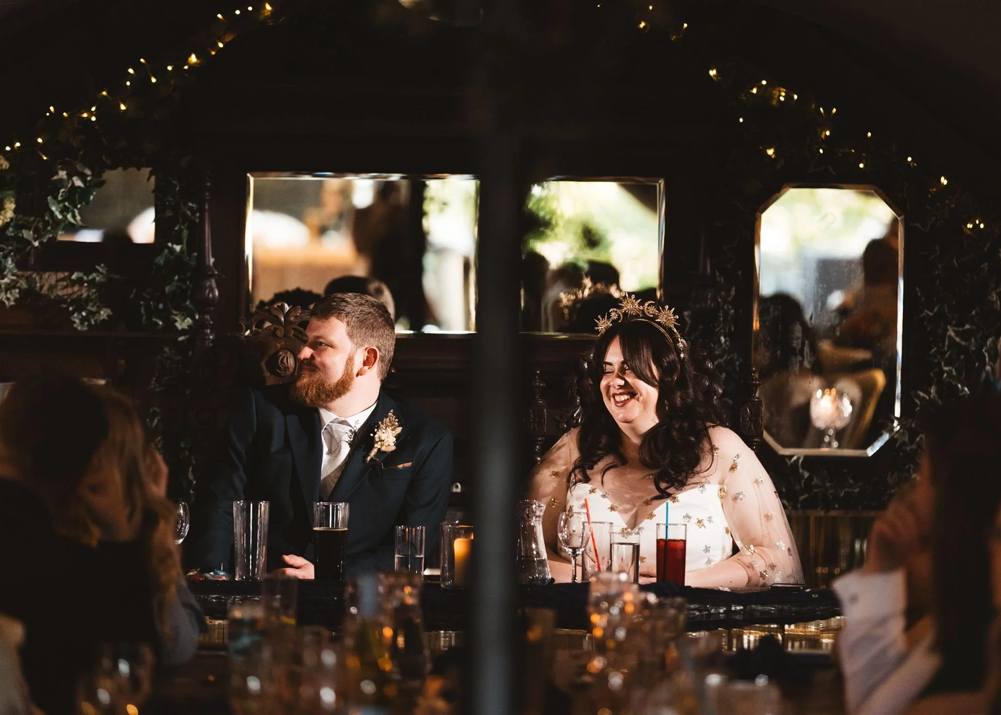 A man with a beard and a woman with long dark hair and a floral crown sit at a table during a wedding reception, smiling and looking to the left. The woman wears a white dress with gold embellishments. There are drinks and glasses on the table, and a