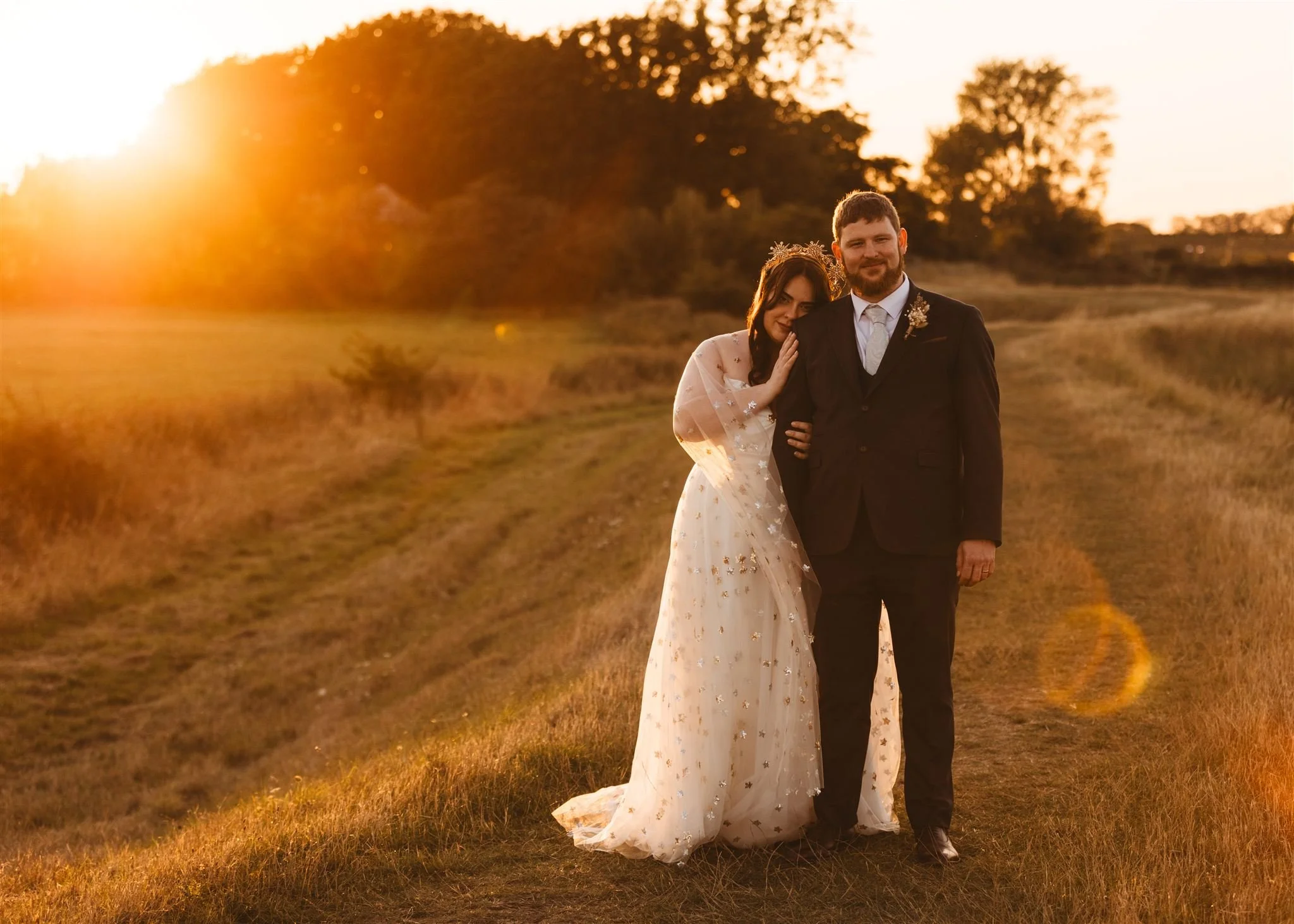 A bride and groom standing together outdoors during sunset, the bride is leaning on the groom's shoulder. The bride is wearing a white wedding dress with floral details, and the groom is in a black suit with a boutonniere. They are on a grassy path w