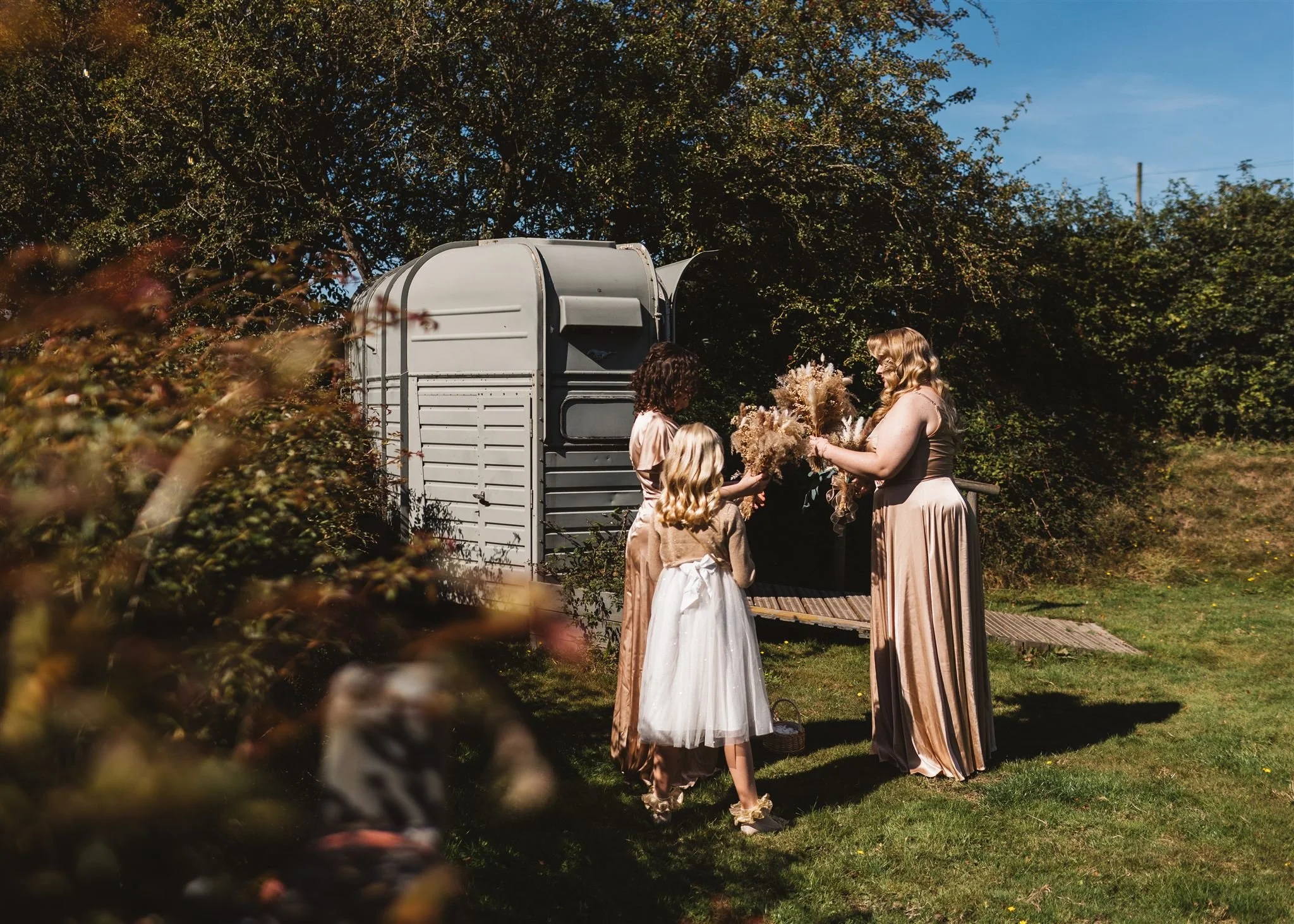 Three women and a child exchanging flowers outdoors on a sunny day, with trees and a wagon in the background.