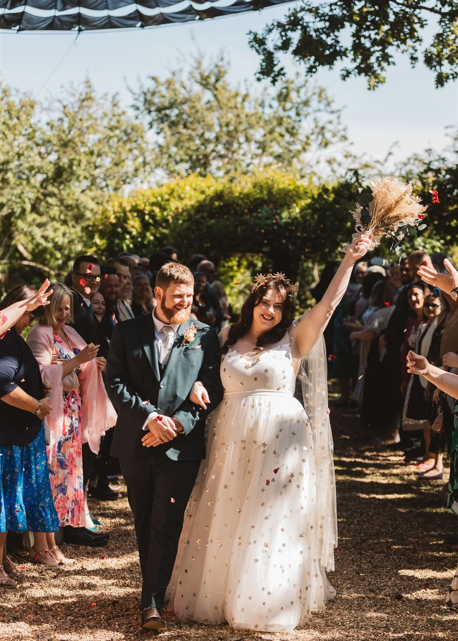 A bride and bridegroom walking together outside at a wedding, with the bride holding a bouquet and smiling, surrounded by guests cheering her on.