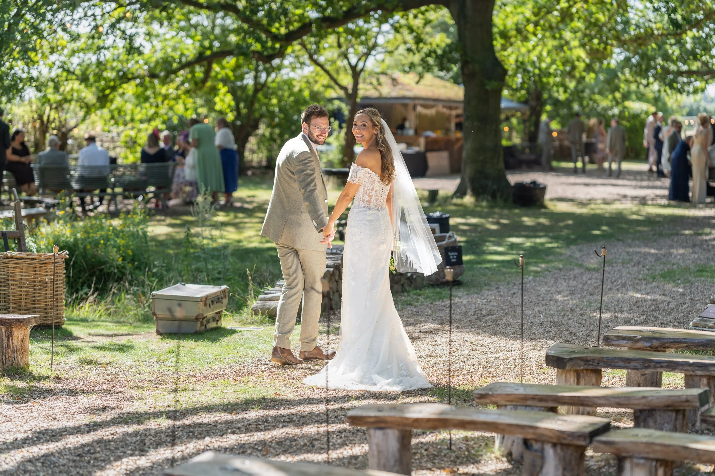 Bride and groom in wedding attire holding hands, smiling, in an outdoor garden setting with guests in the background.