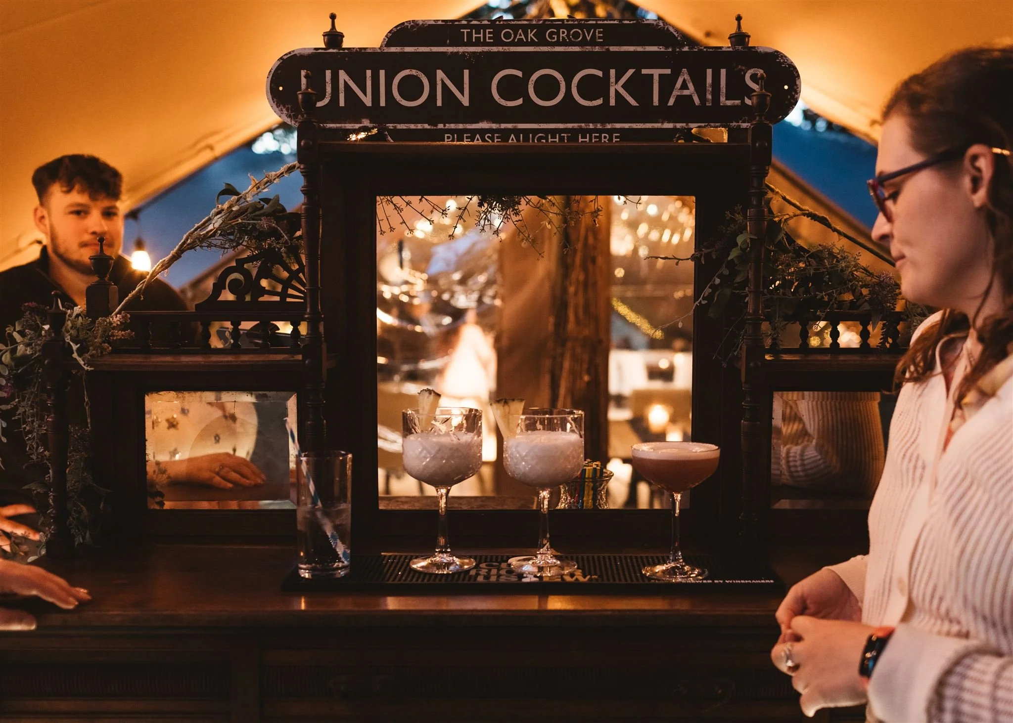A woman standing at a bar counter with three colorful cocktails in glasses. Behind the bar, a bartender is visible, and a sign above reads 'Union Cocktails.' The setting appears to be a cozy outdoor tent with warm lighting.