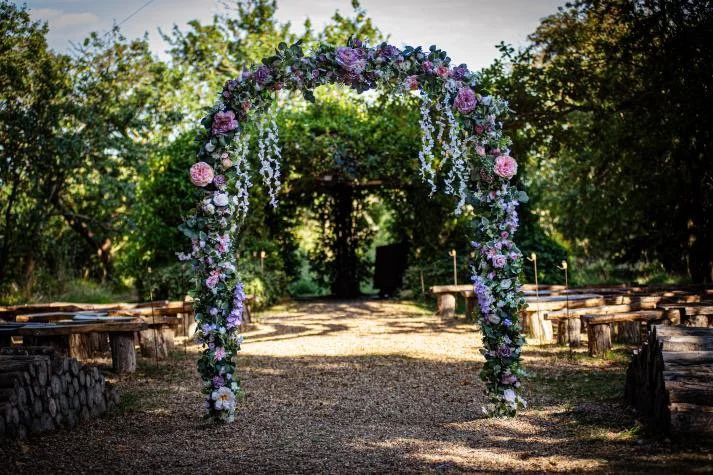 A floral arch decorated with pink, purple, and white flowers, set up outdoors on a gravel pathway surrounded by trees, for a wedding ceremony.