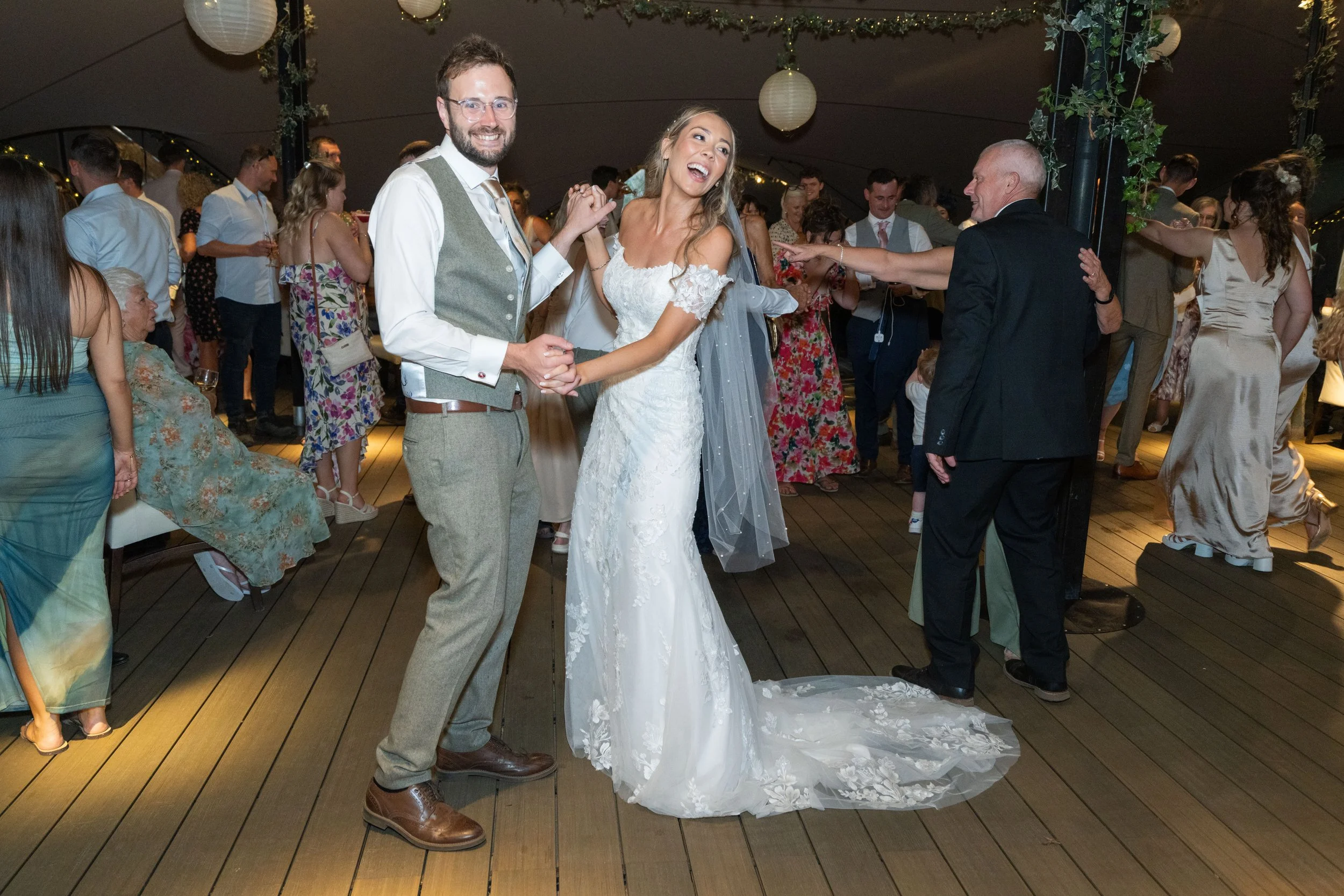 A bride and groom dancing at their wedding reception with guests around them, some taking photos and others dancing. The bride is in a white lace wedding dress with a veil, and the groom is in a gray vest and dress pants. The scene is under a decorat