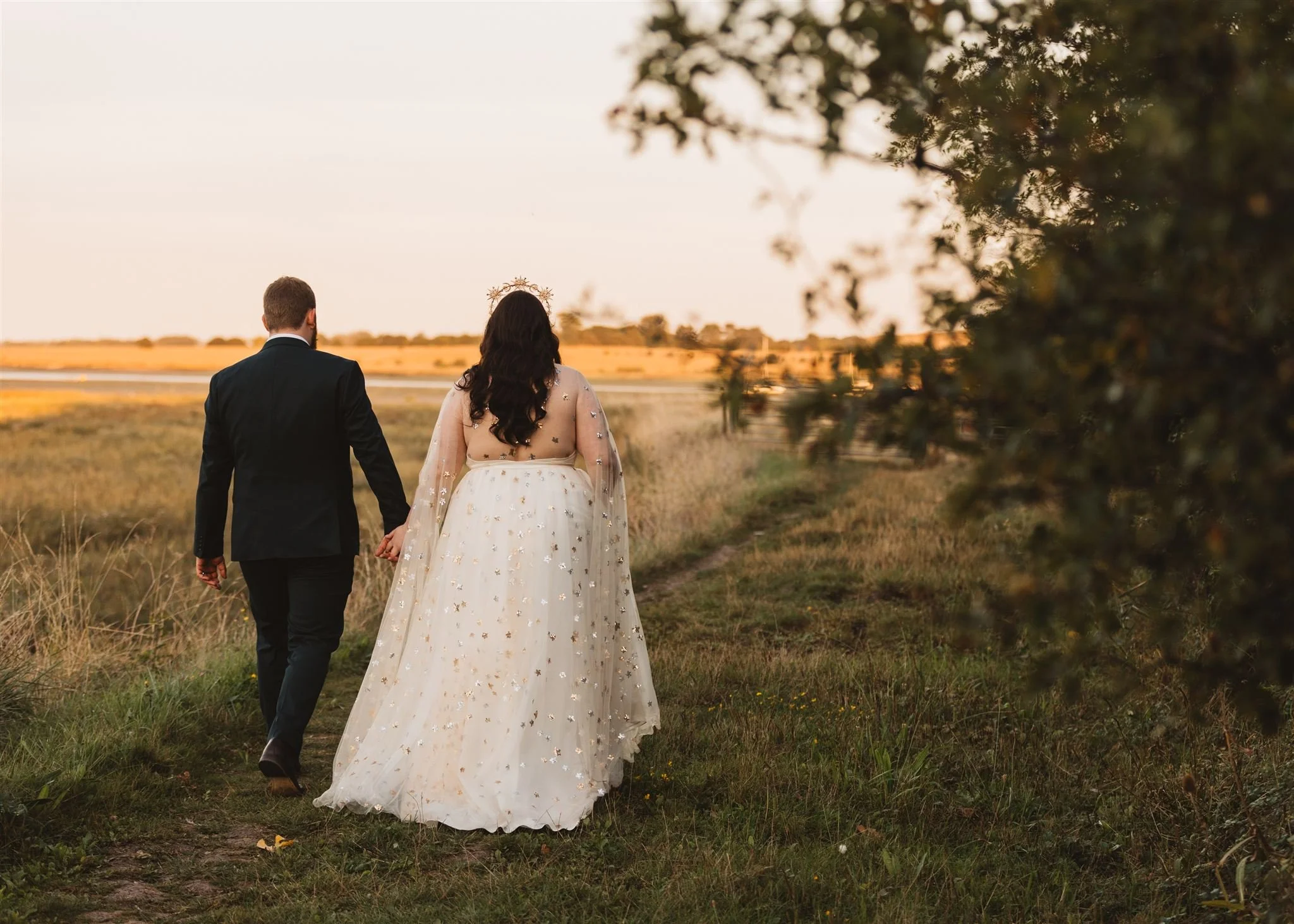 A bride and groom walking hand in hand on a grassy path during sunset in a field, with the bride wearing a white dress and tiara and the groom in a black suit.