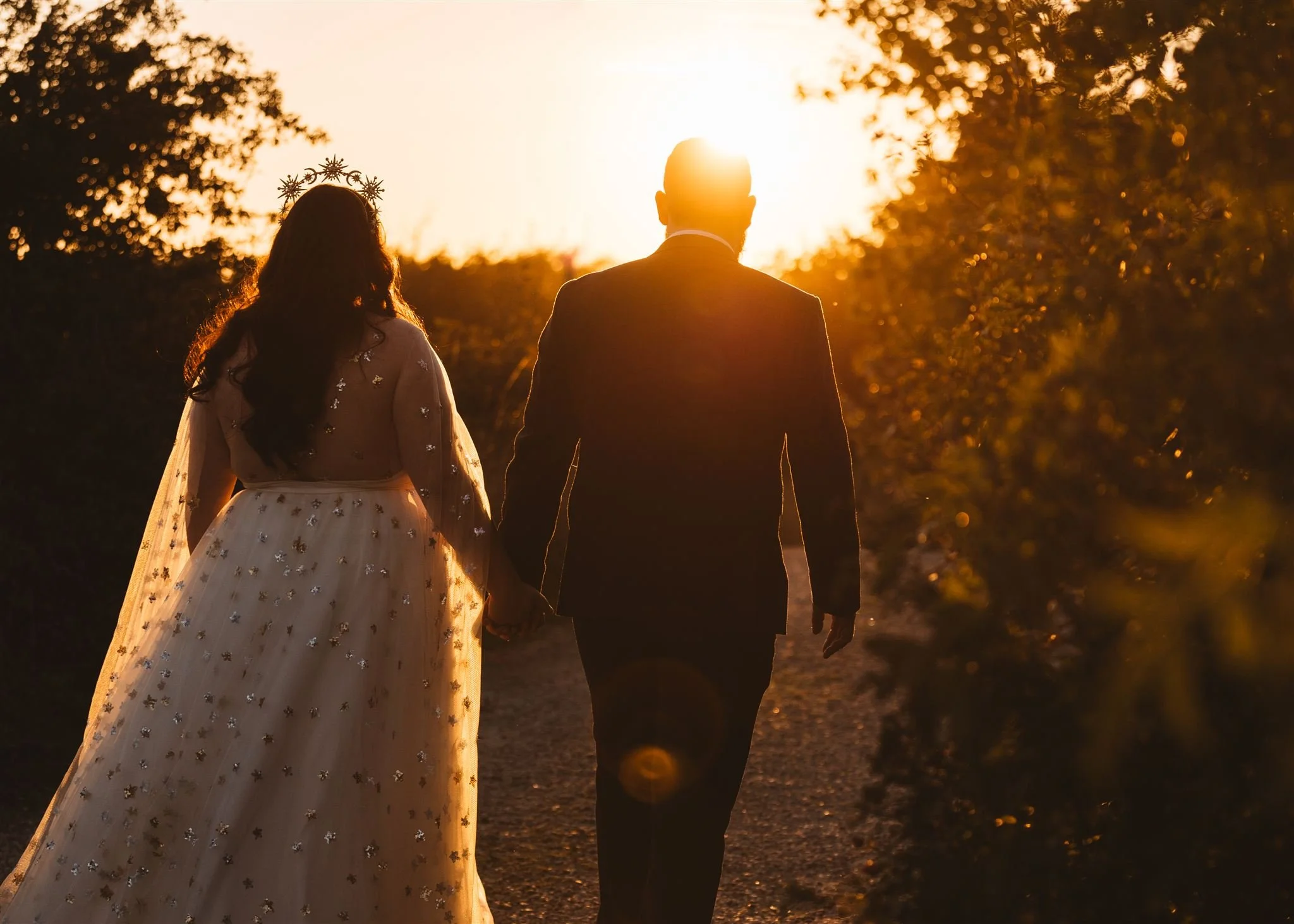 A couple holding hands walking outdoors at sunset, with the woman's dress and a crown, and sunlight creating a silhouette effect.