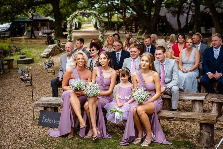 Group of wedding guests seated outdoors, including bridesmaids in lavender dresses and children holding bouquets, with a wedding arch decorated with flowers in the background.