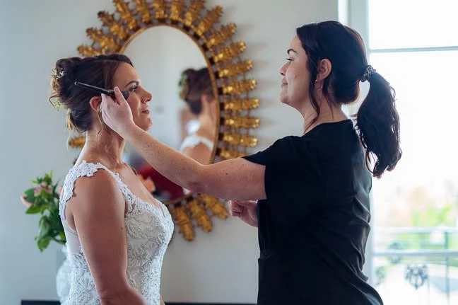 Makeup artist applies makeup to bride in a room with a decorative mirror and a window with natural light.