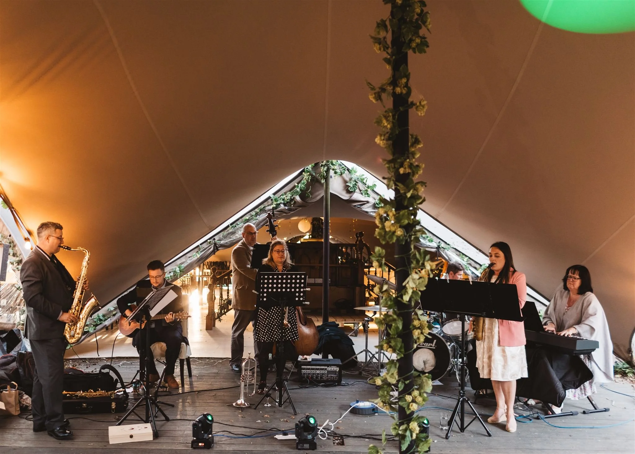 A jazz band performing on stage inside a tent with a vaulted ceiling, decorated with greenery and flowers. The band includes a saxophonist, a guitarist, a bassist, a pianist, a drummer, and a vocalist, all of whom are engaged in their performance.