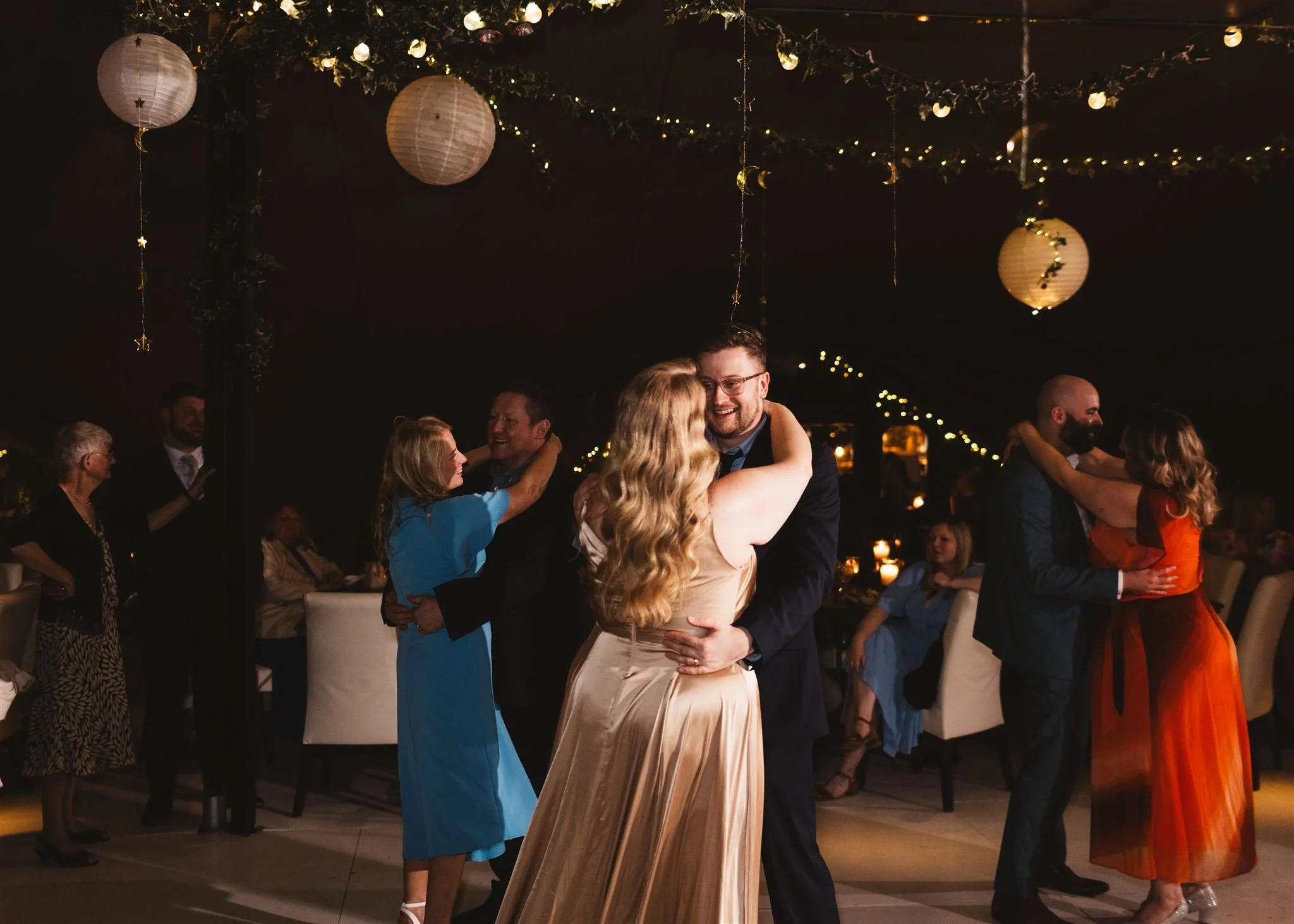 Couples dancing at a wedding reception with moon and star paper lanterns and string lights hanging from the ceiling.