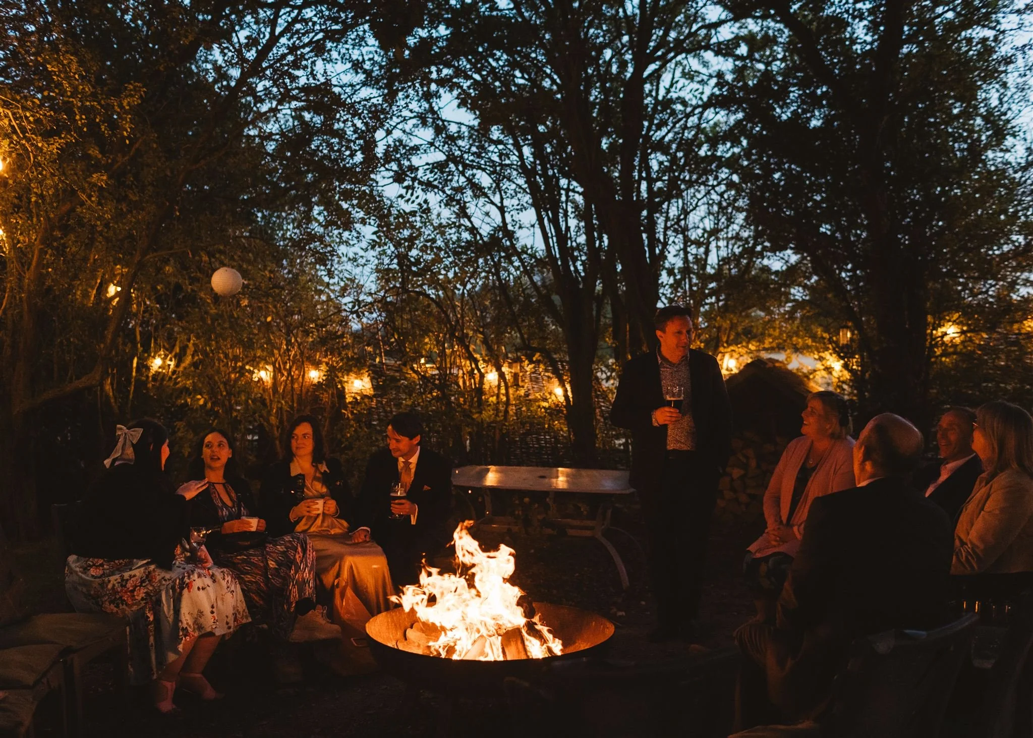 People gathered around a fire pit outdoors at dusk, some sitting and some standing, enjoying drinks in a wooded area with string lights hanging in the trees.