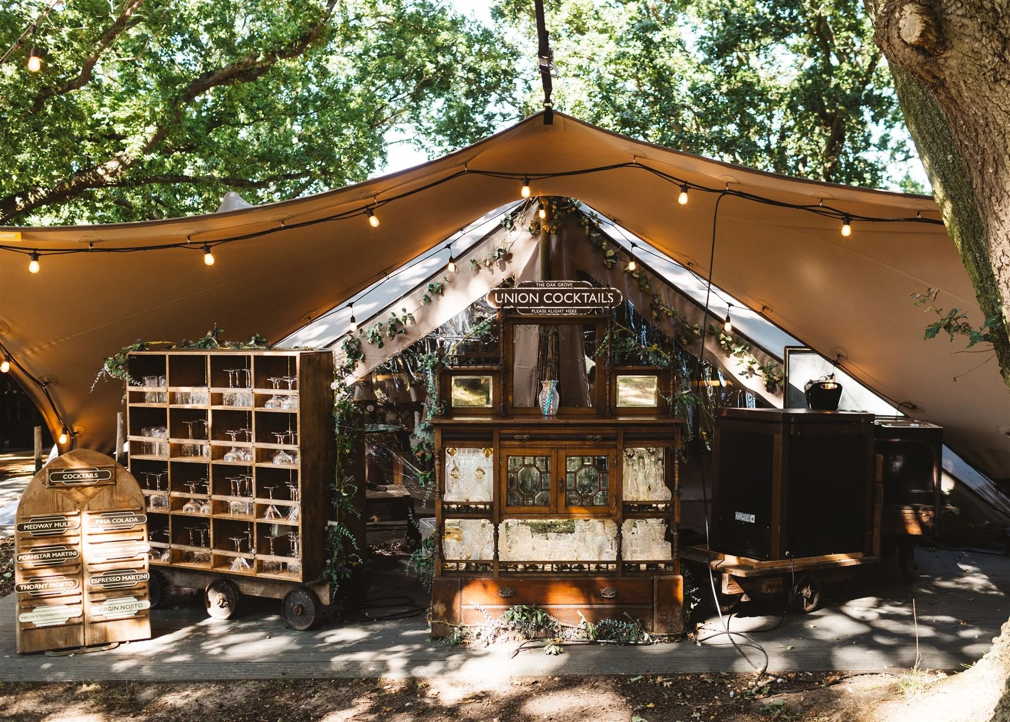 An outdoor bar setup under a tent with string lights, greenery, and a sign reading 'Union Cocktails'.