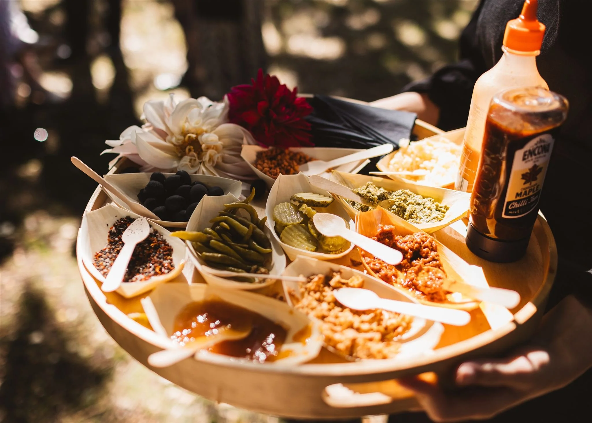 A wooden tray holding small bowls of various pickled vegetables, sauces, and condiments, with a bottle of barbecue sauce on the side, and some flowers in the background.