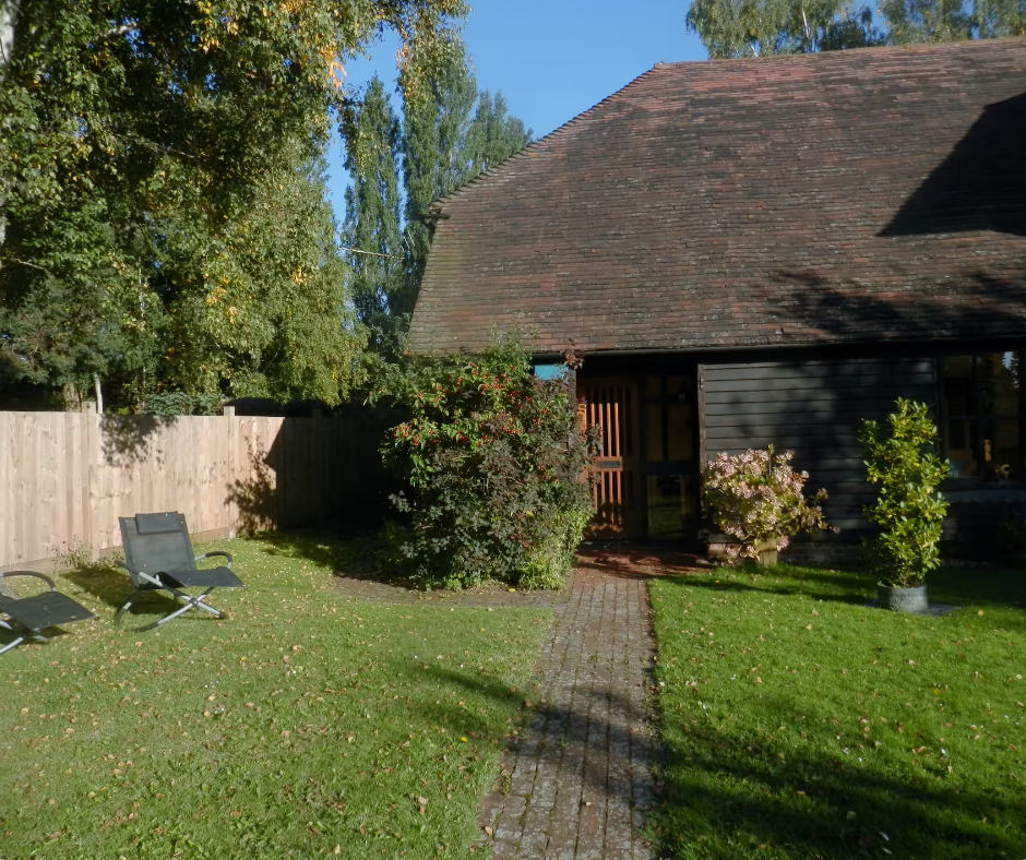 A backyard garden with green grass, seating chairs, flowering bushes, a brick path, and a black wooden house with a tiled roof under a clear blue sky.