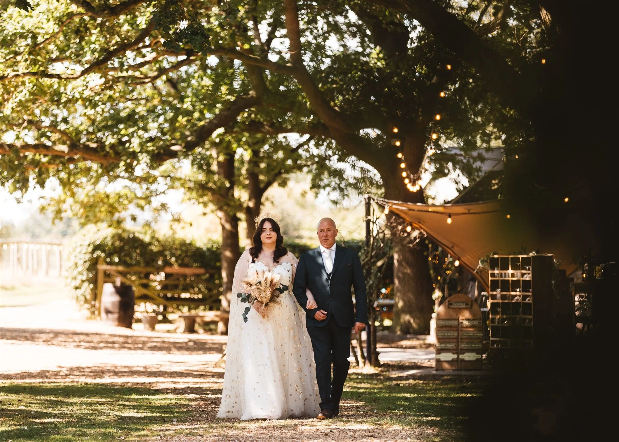 A bride and an older man walking down an outdoor garden aisle, with trees and string lights overhead, during what appears to be a wedding ceremony.