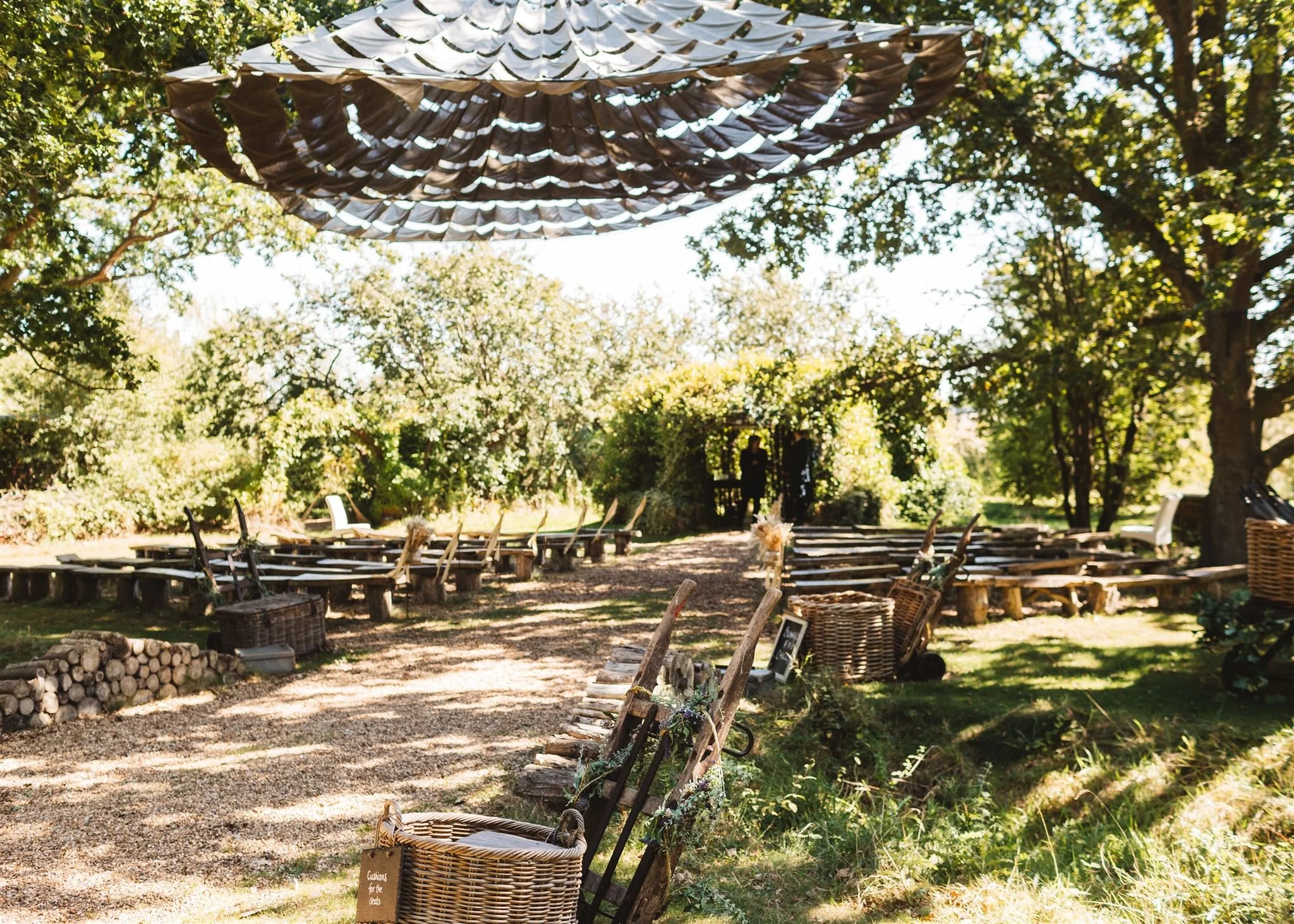 Outdoor wedding setup with wooden benches and chairs under a canopy, surrounded by trees and greenery.
