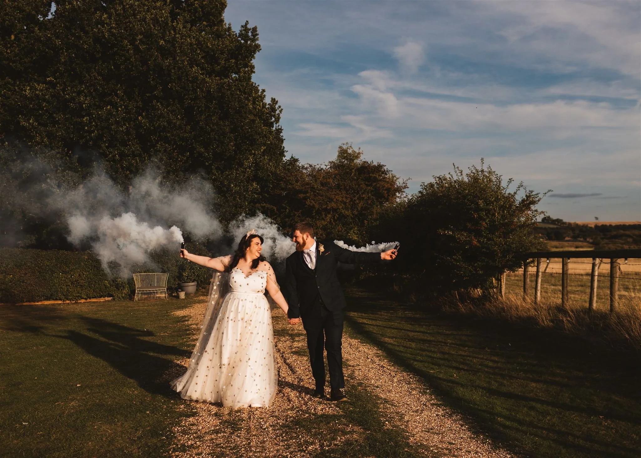 A bride and groom walking hand in hand outdoors at sunset, holding smoke grenades and smiling at each other.
