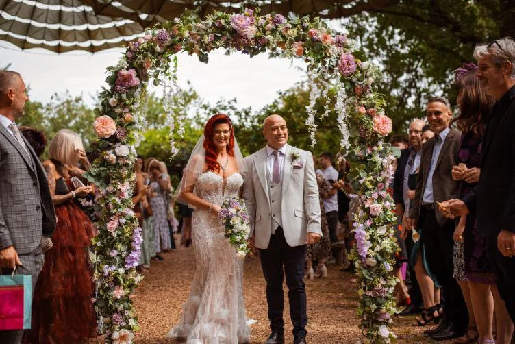 Bride and groom walking under a floral arch during their outdoor wedding ceremony, surrounded by guests.