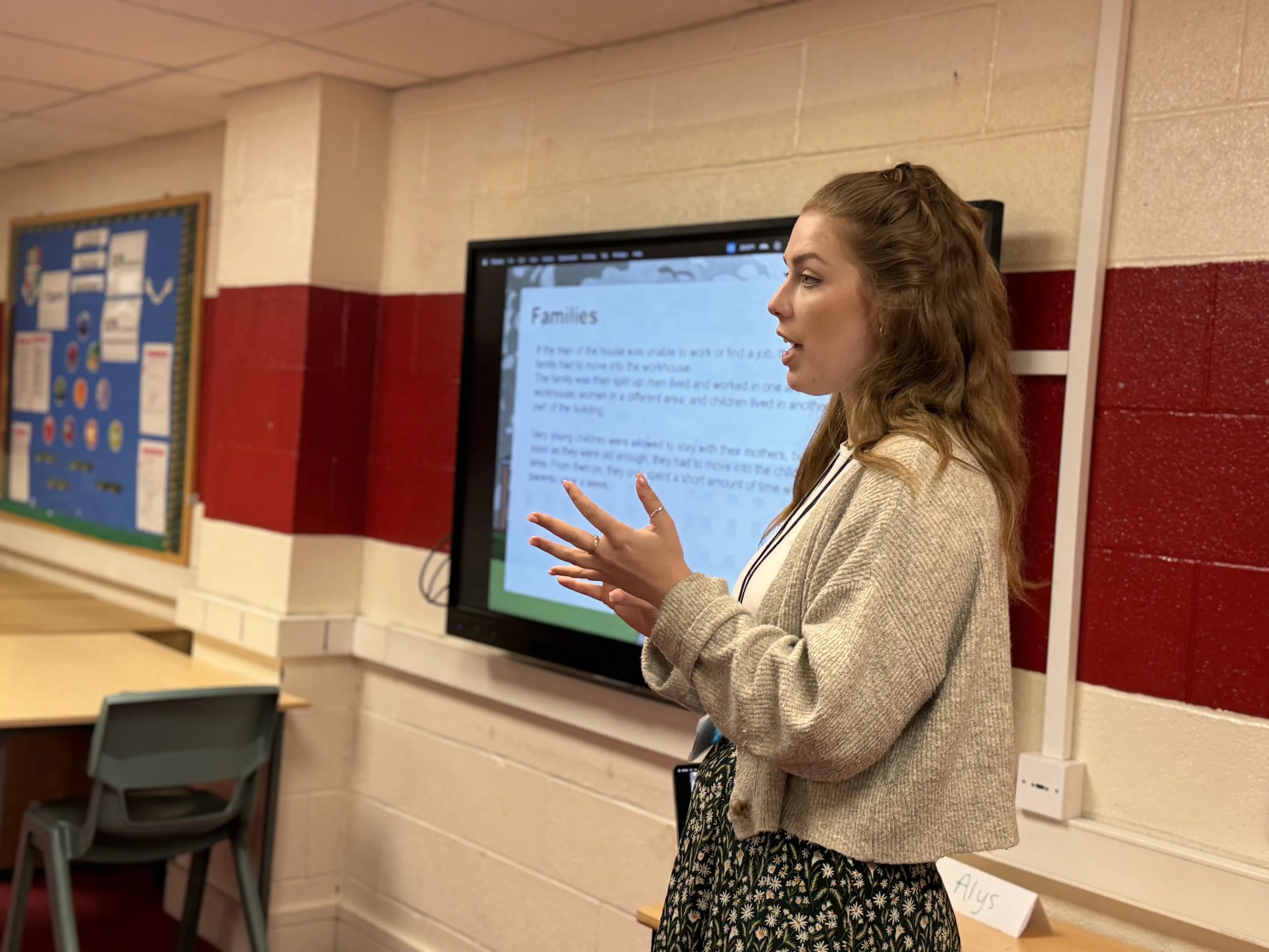 A woman standing in front of a digital display, presenting to a classroom or group. She is gesturing with her right hand and has long, wavy hair. She is wearing a beige cardigan over a patterned dress. The background includes a blue bulletin board on the wall and a sign labeled "Alys" on the table.