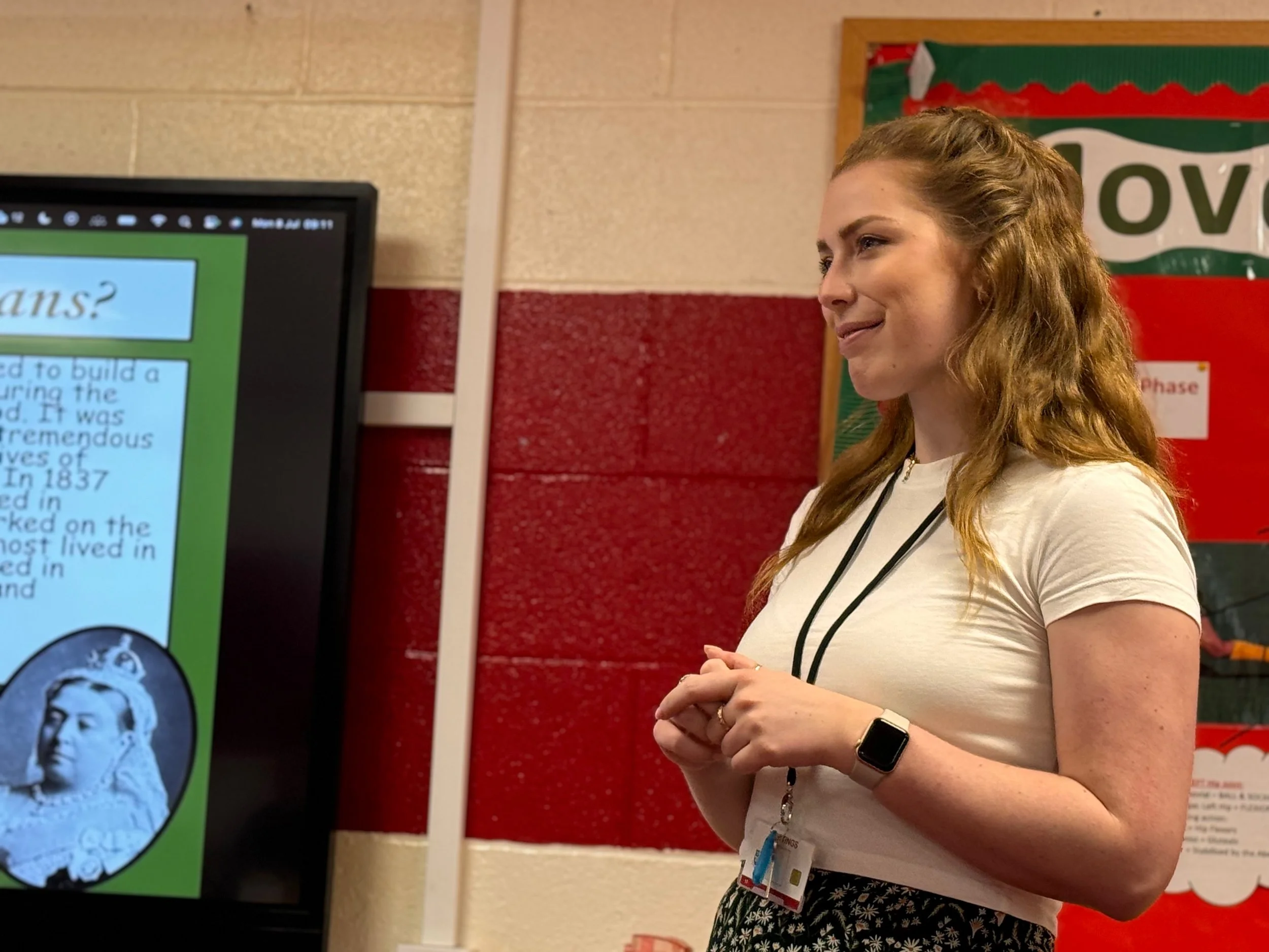 A young woman with wavy, reddish-blonde hair standing in front of a presentation display, smiling and wearing a white T-shirt, a smartwatch, and an ID badge around her neck.