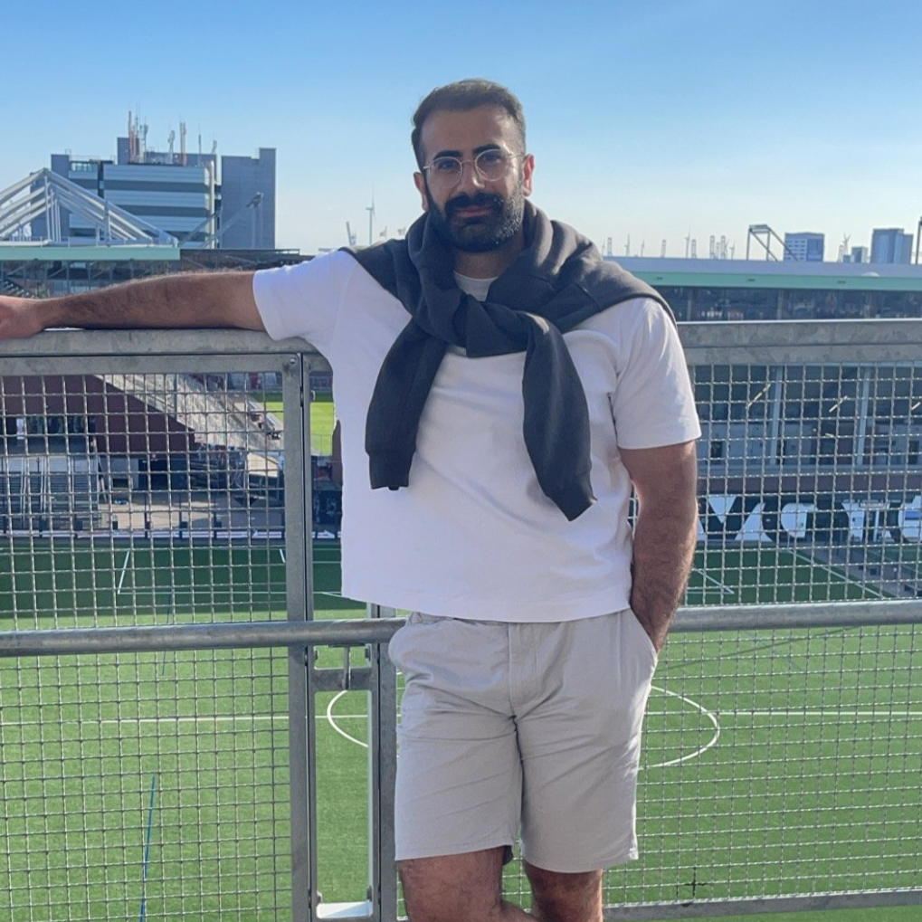 Mann mit dunklem Bart, Brille und hellen Shorts på dem Balkon eines Fußballstadions, Blick auf das Spielfeld und die Stadt im Hintergrund.