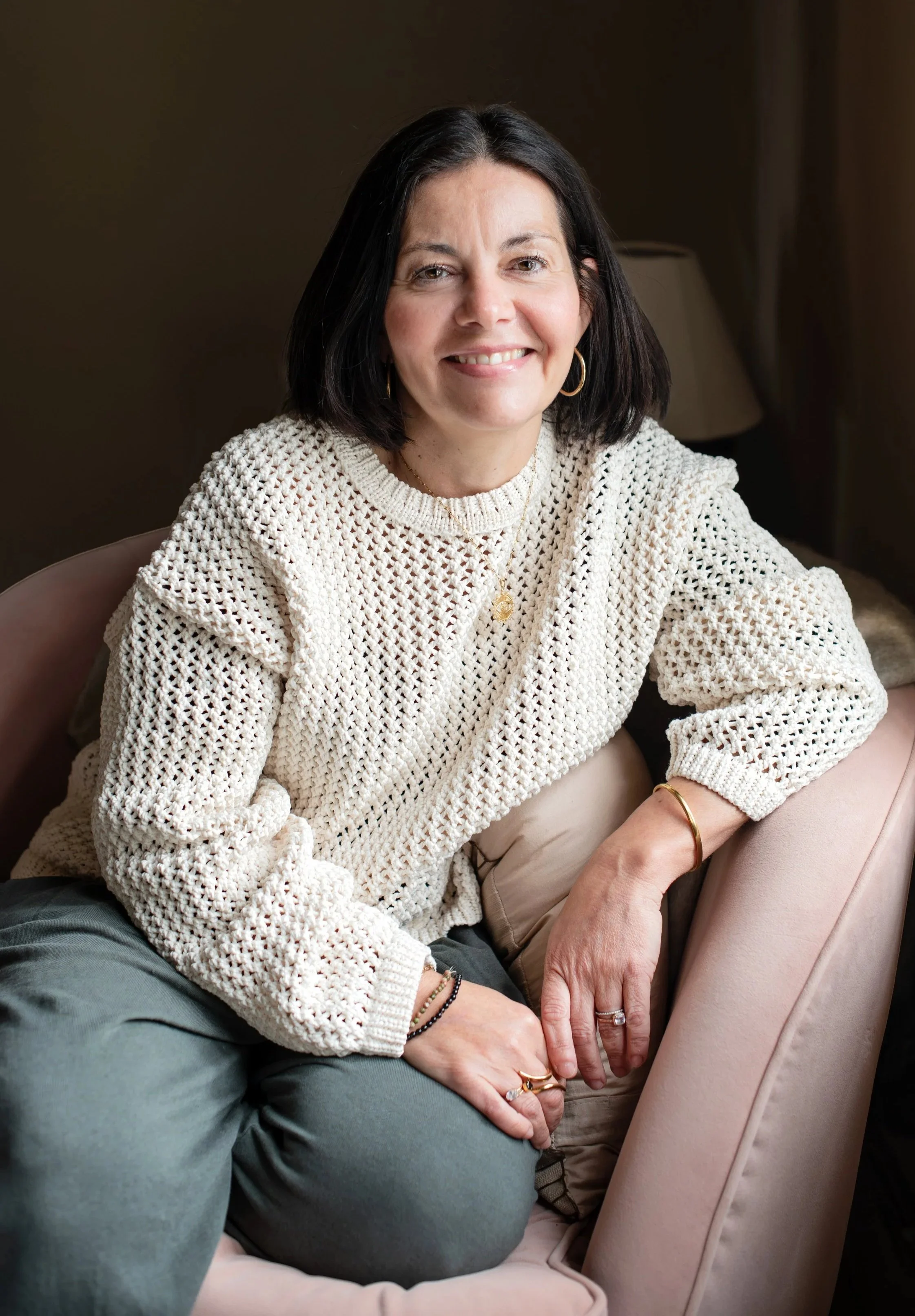 A woman with dark hair, wearing a cream-colored crochet sweater, gold hoop earrings, and jewelry, smiling while sitting on a beige sofa, with another person's hand touching her knee.