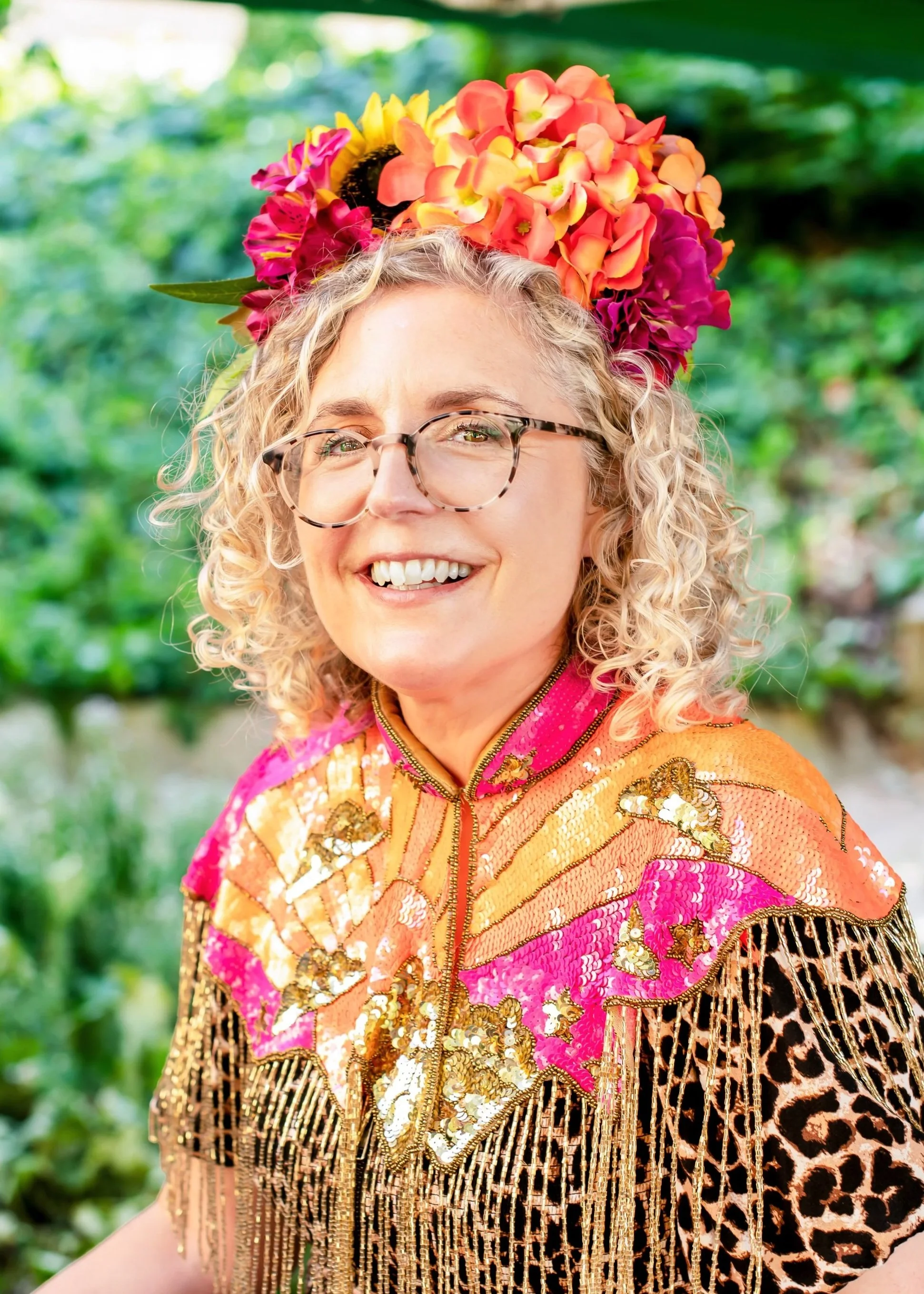 A woman with curly blonde hair and glasses wearing a vibrant, colorful traditional outfit with gold fringes and floral embroidery, standing outdoors with green foliage in the background. She is smiling and has a headpiece made of pink, yellow, and orange flowers.