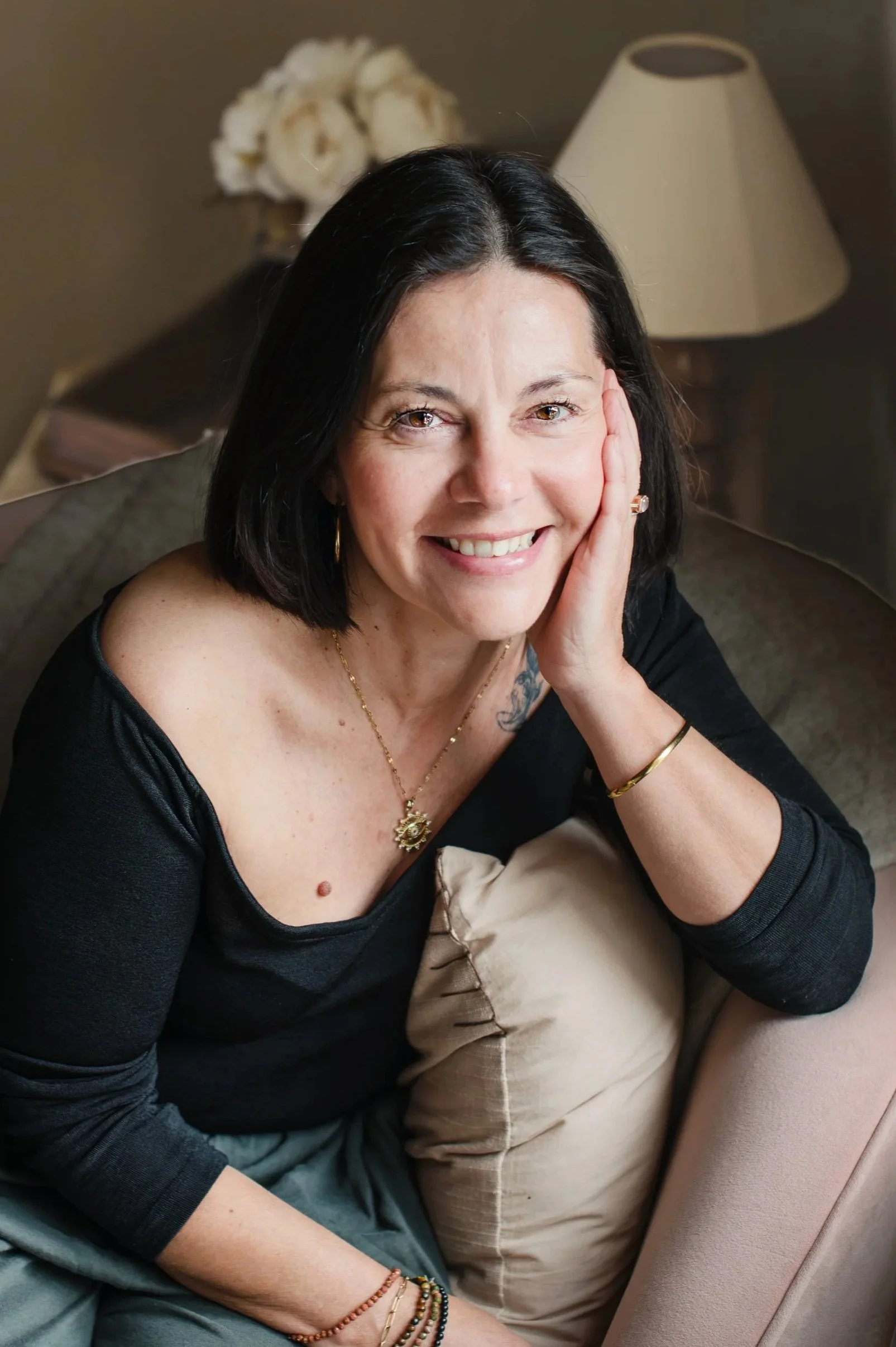 A kundalini yoga practitioner with black hair smiling, resting her head on her hand, sitting on a beige couch, with a pillow and a lamp in the background. Shot by Wellness Brand Photographer, Emily Dyer Creative