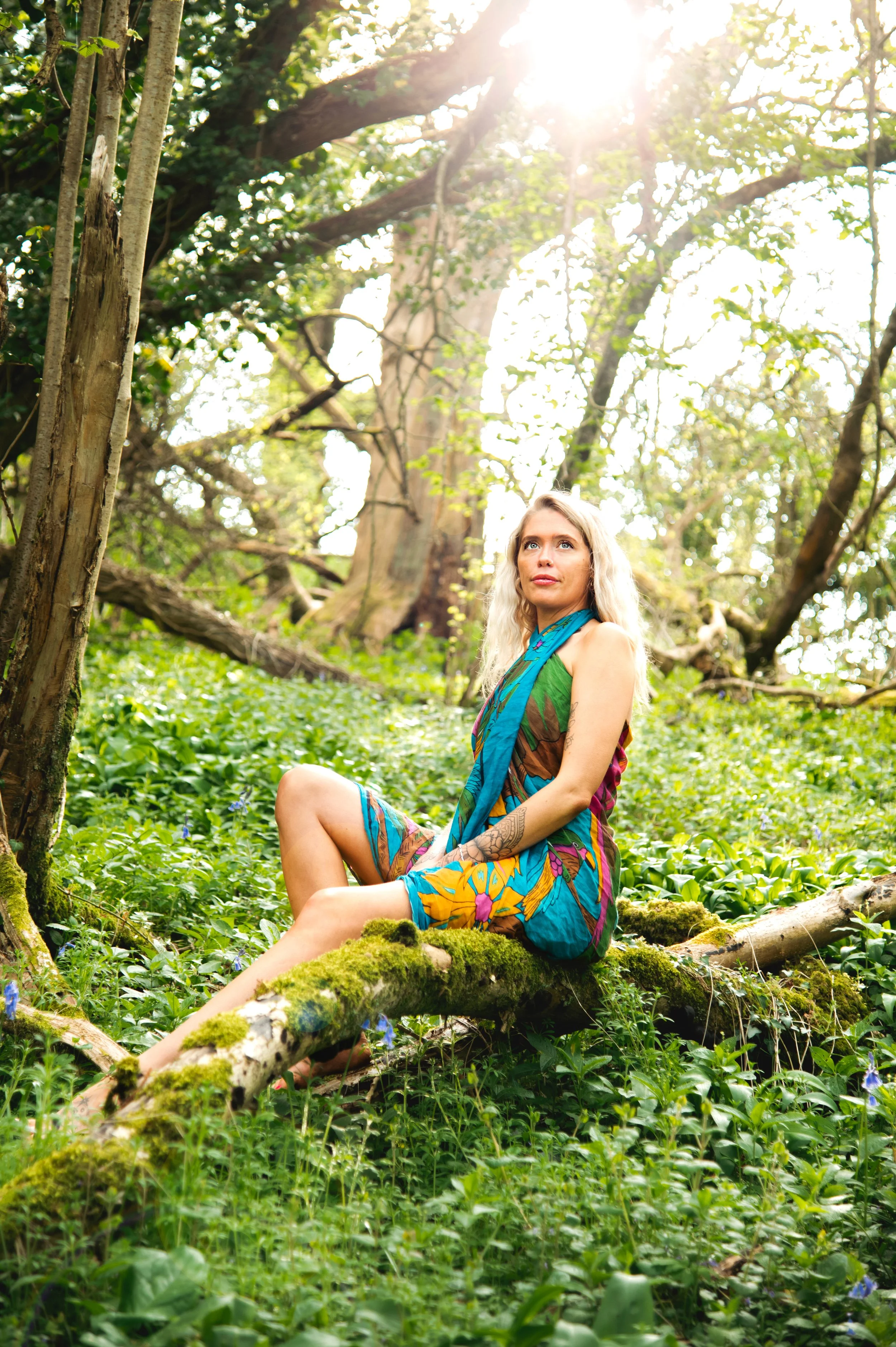 A female breathwork practitioner with long blonde hair sitting on a mossy fallen tree trunk in a lush green forest, wearing a colorful, sleeveless dress with a floral pattern, looking upward with a serene expression. Shot by Emily Dyer Creative