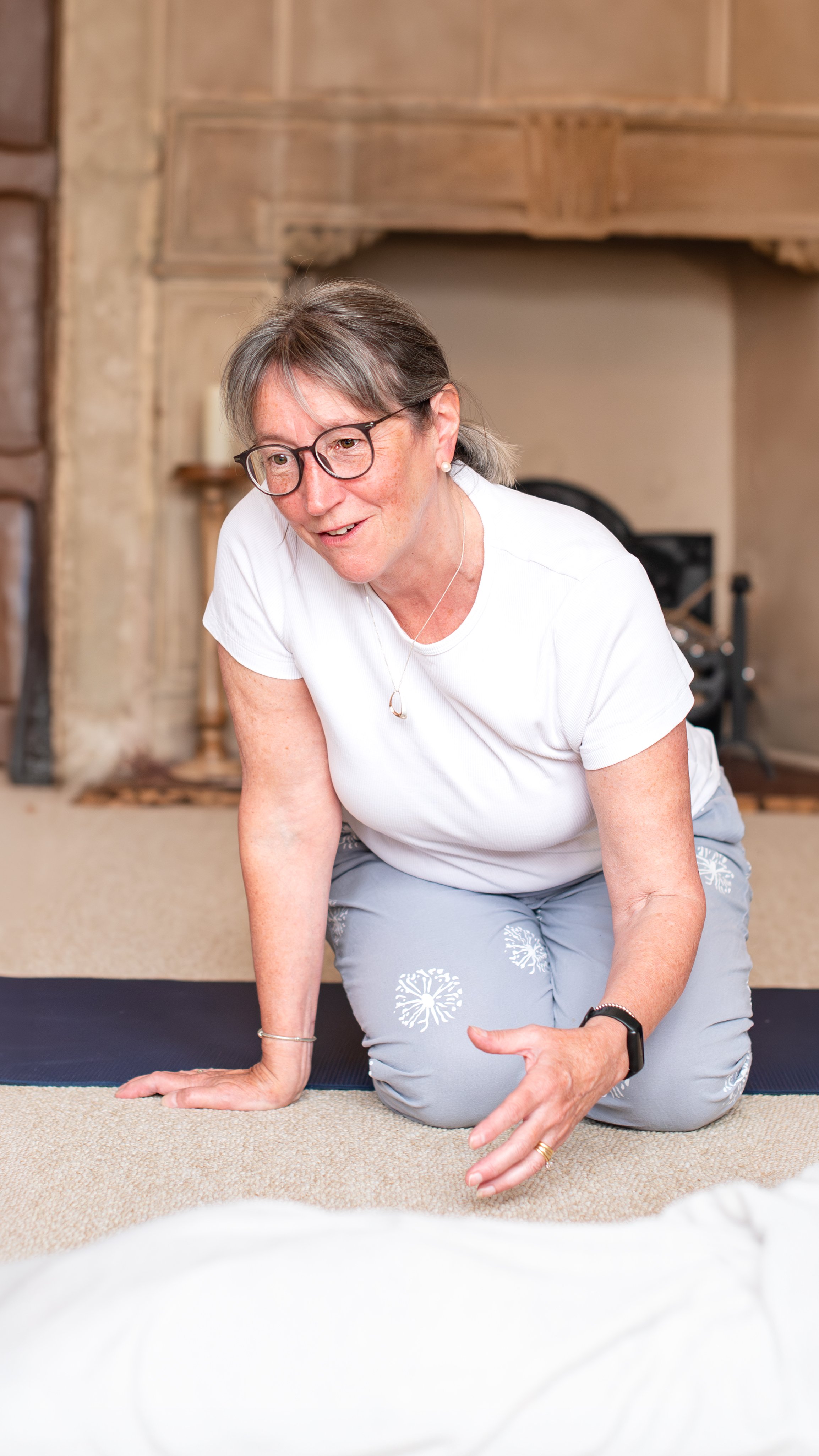 A woman with glasses and gray hair performing a yoga therapy on a carpeted floor, in front of a fireplace. Shot by Wellness Brand Photographer, Emily Dyer Creative