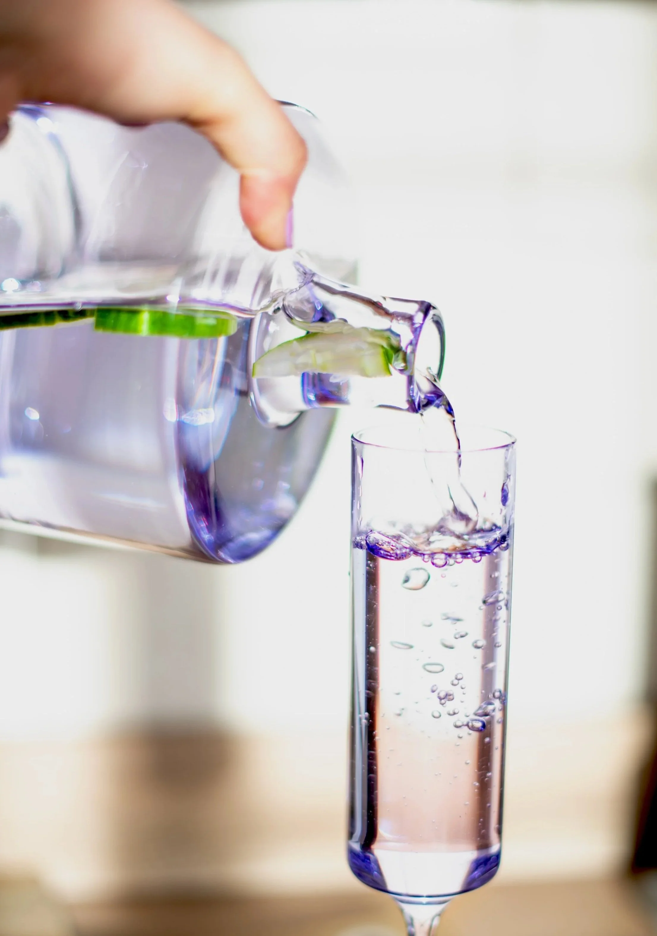 Person pouring water from a lilac glass pitcher into a tall, lilac glass. The water is lilac-tinted, and cucumber slices are visible inside the pitcher. The background is blurred and neutral. Shot in Witney, Oxfordshire, Wellness Brand Photographer