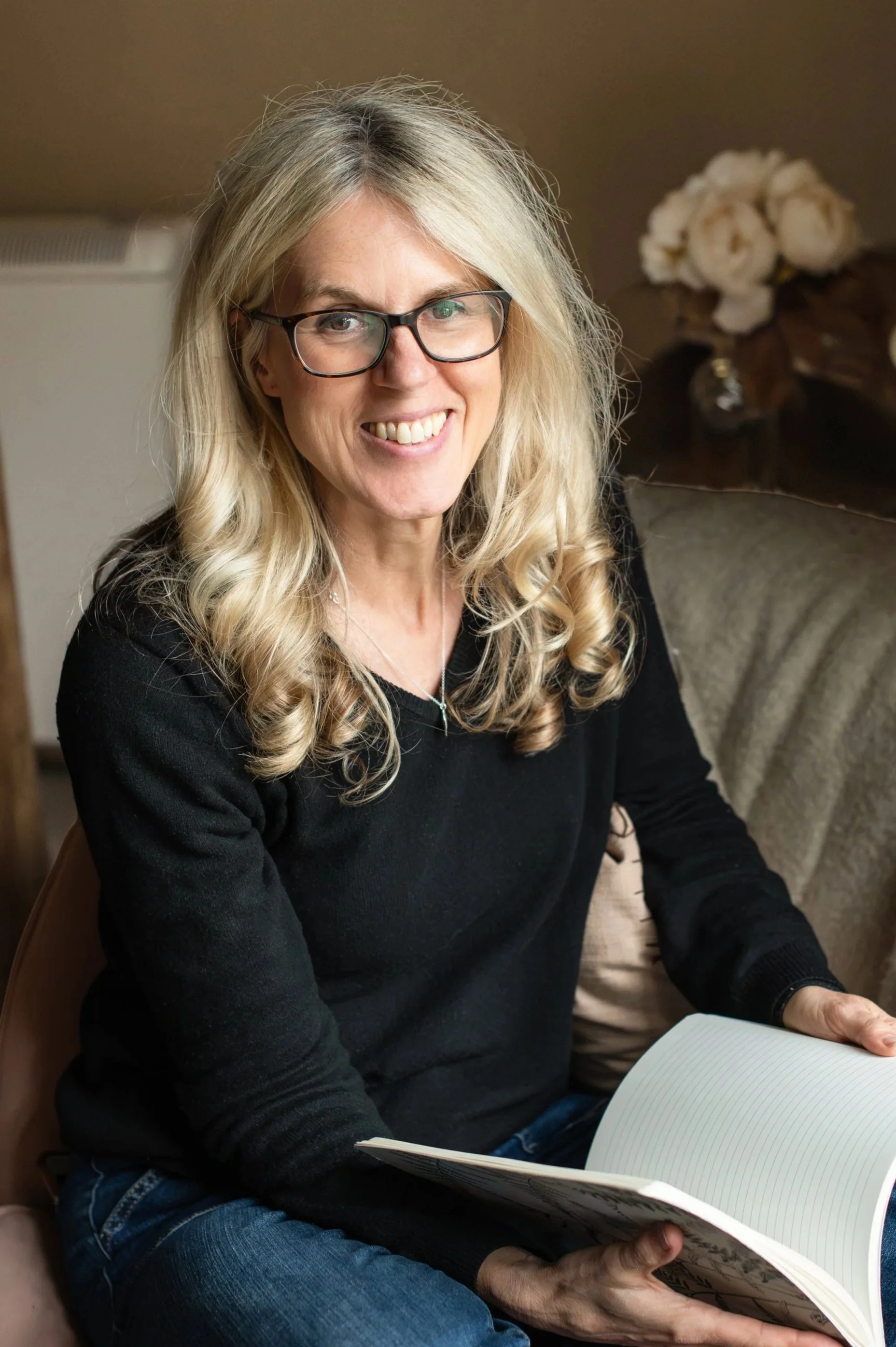 A Yoga Nidra teacher with blonde hair, glasses, and a black top sitting indoors, holding an open notebook or journal with lined pages. She is smiling. Shot by Wellness Brand Photographer, Emily Dyer Creative