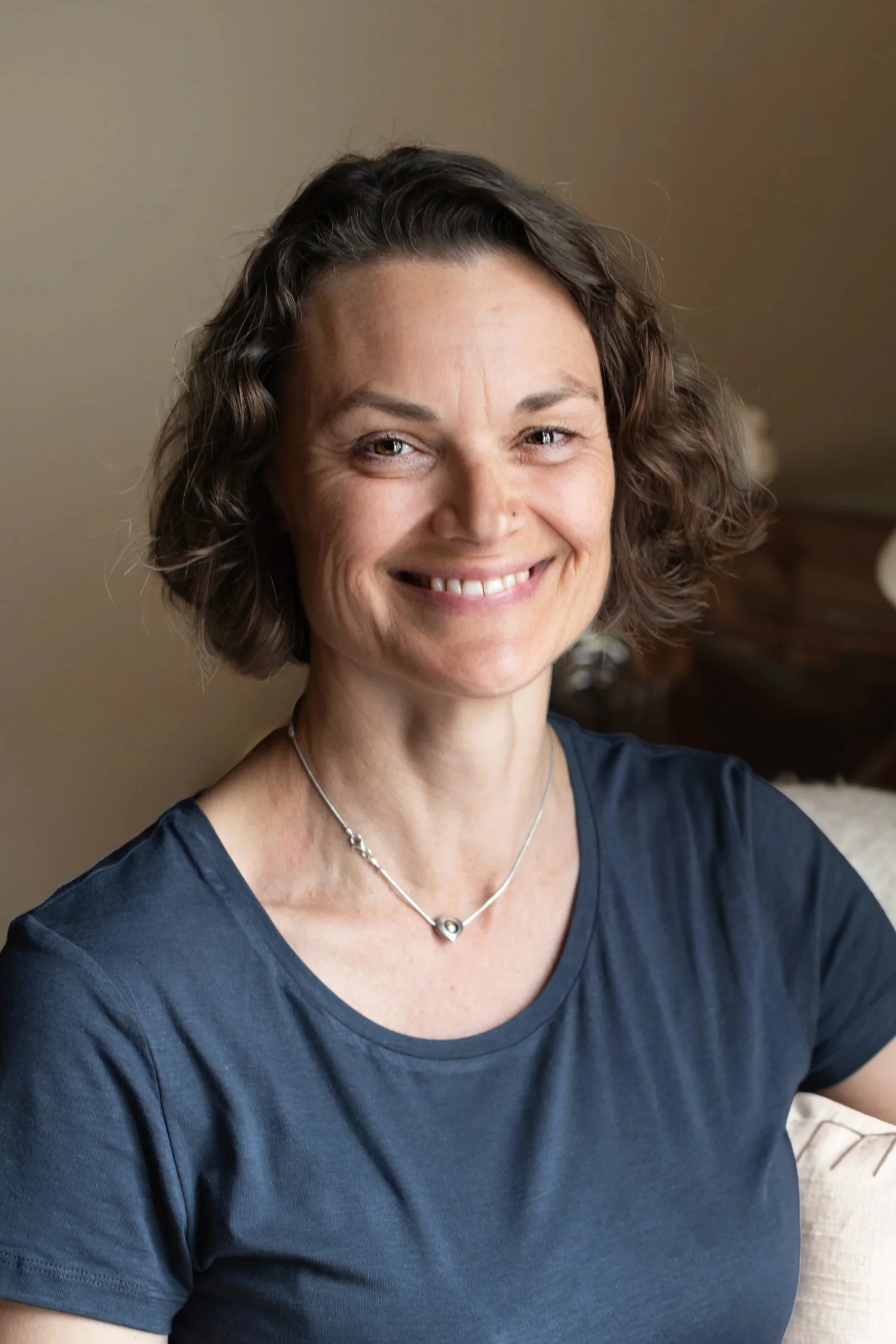 A smiling woman with wavy short brown hair, wearing a dark blue top and a delicate silver necklace, sitting in a cozy indoor setting.