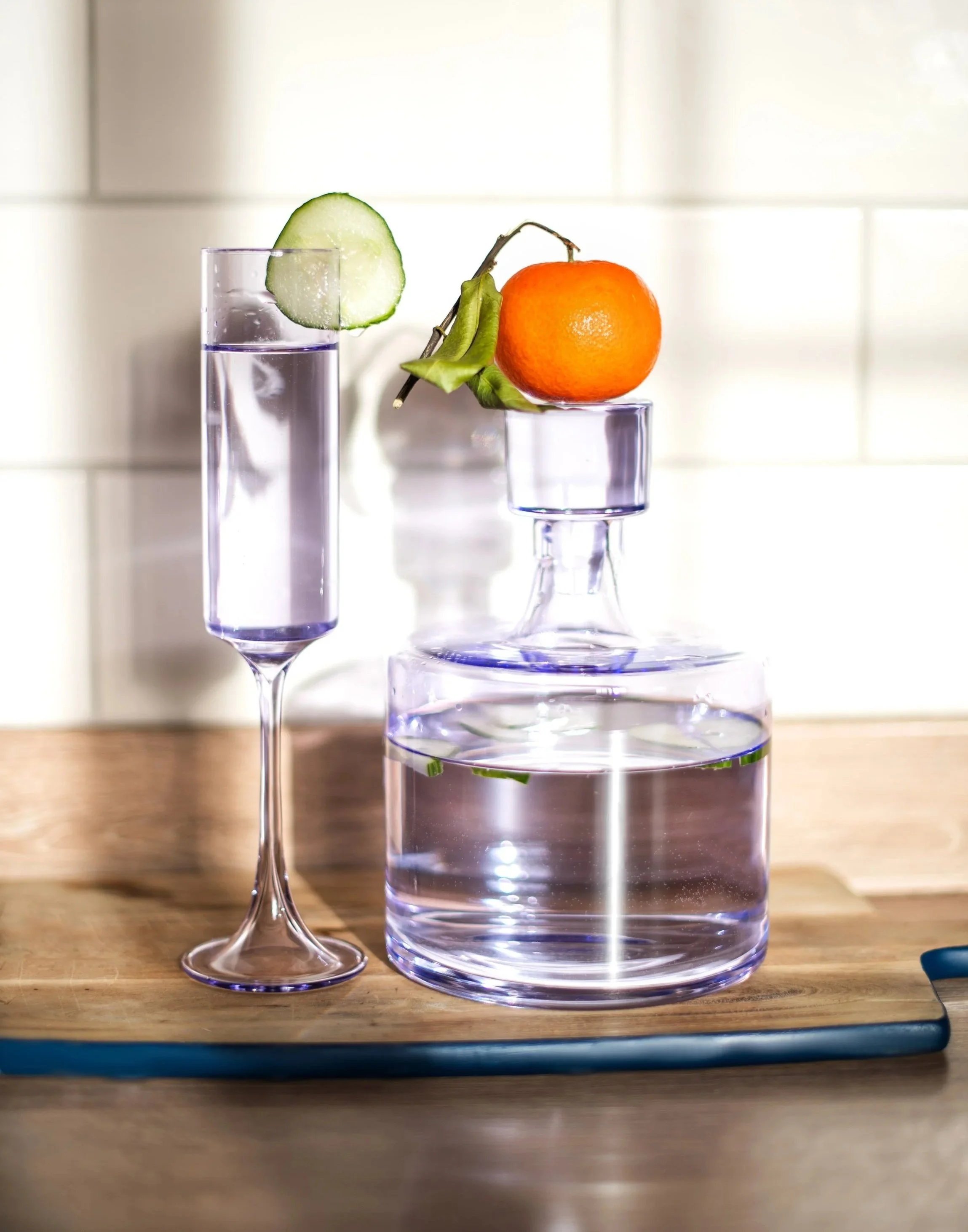 A clear glass with purple liquid and a slice of cucumber on its rim, a small glass with an orange, and a small glass with a green leaf, on a wooden surface with a tiled wall background. Shot by Wellness Brand Photographer, Emily Dyer Creative