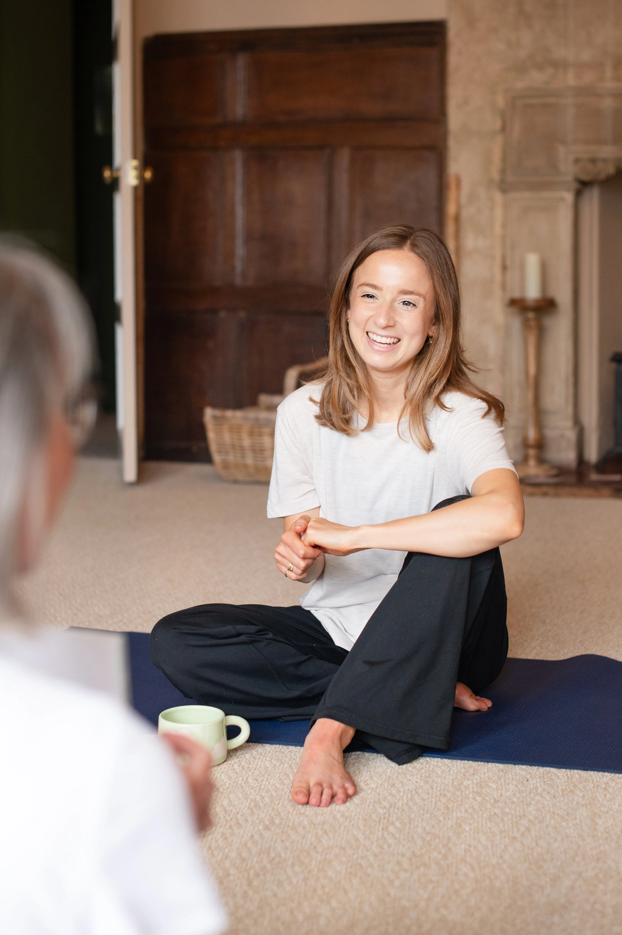 A young woman with shoulder-length light brown hair sitting cross-legged on a yoga mat, smiling, in a room with a beige carpet, a wooden cabinet, and a candle holder. She is wearing a white t-shirt and black pants, and appears to be engaged in a conversation with a person partially visible in the foreground, holding a mug.