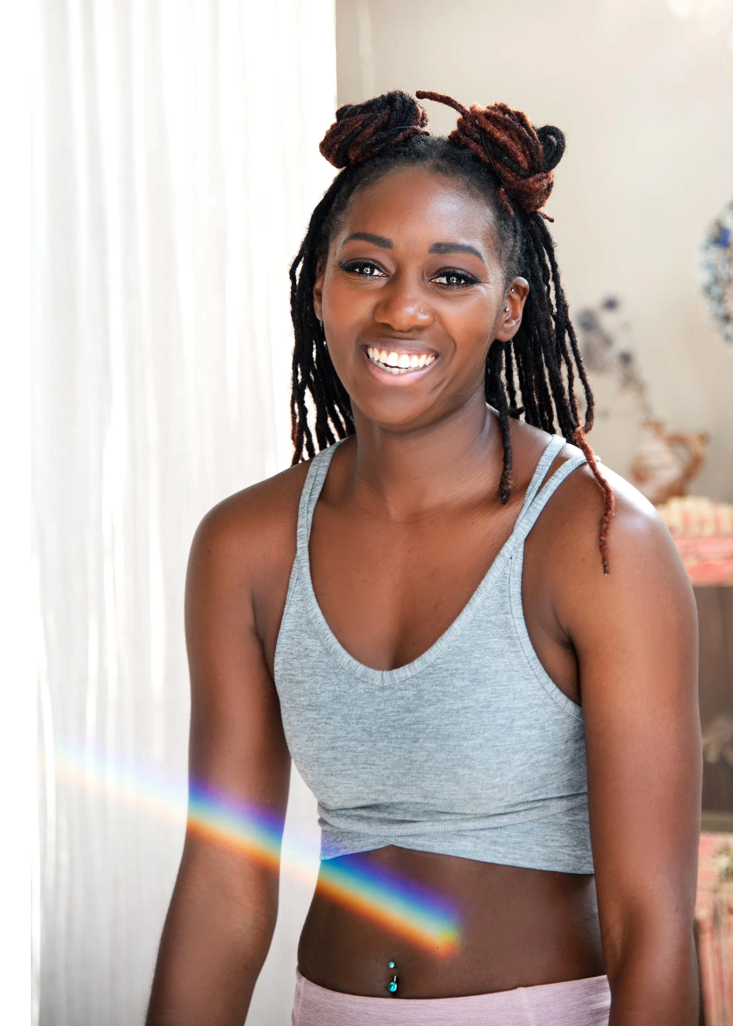 A smiling massage therapist woman with dreadlocks wearing a gray tank top and pink shorts, standing indoors with a rainbow light effect across her body and a blurred background.