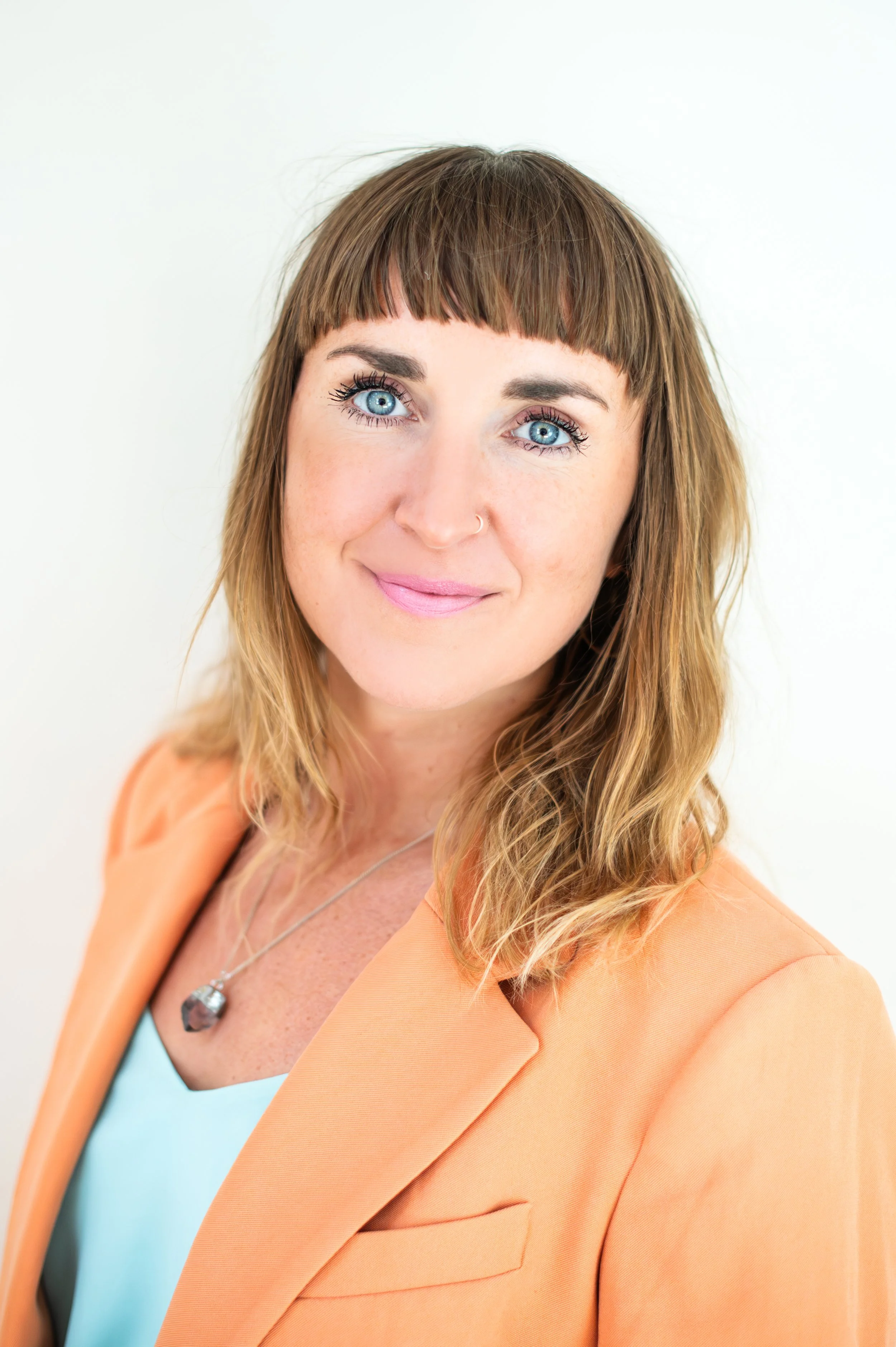 A woman with light brown hair, blue eyes, and a nose piercing, wearing a light blue top and an orange blazer, smiling against a white background.