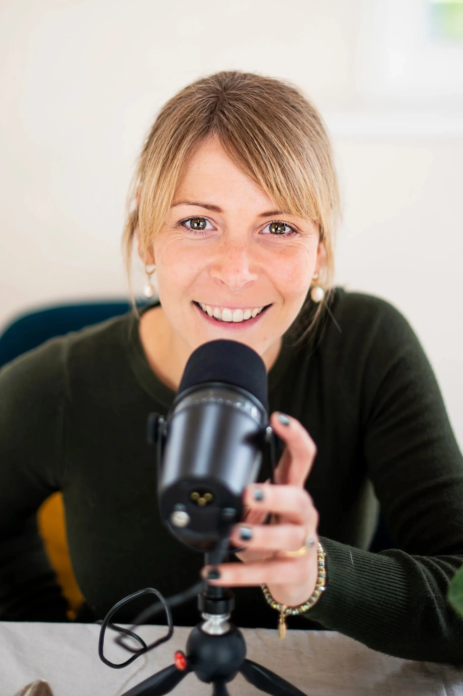 A woman with light brown hair and brown eyes smiling at the camera while holding a camera and sitting at a desk.